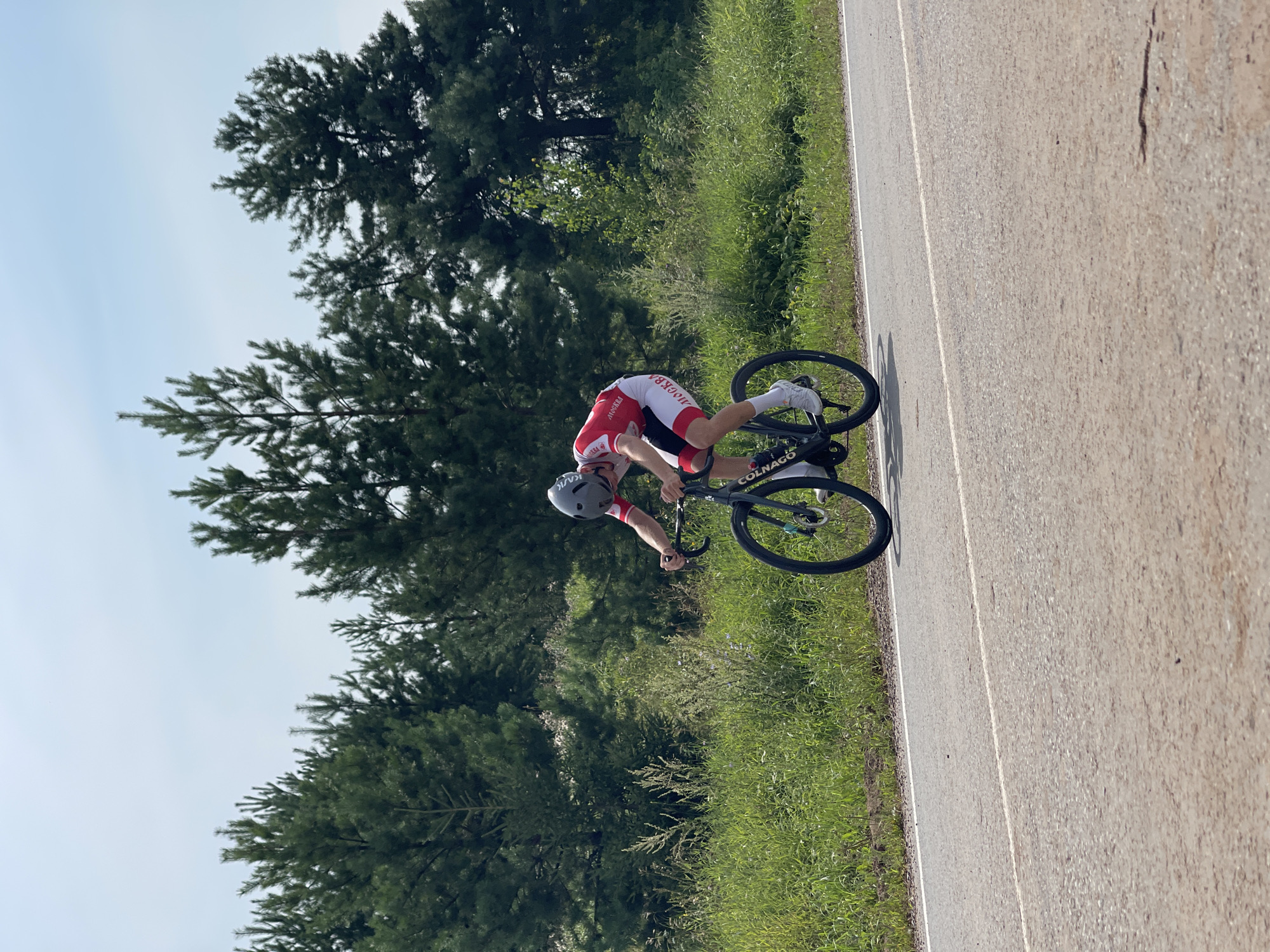 A male cyclist wearing a red and white racing suit and a grey helmet rides a black Colnago road bike on an asphalt road. He is wearing white socks and cycling shoes. The cyclist is positioned in the middle ground of the image, moving from left to right. Behind him, on the left side of the road, is a stretch of lush green grass and vegetation with scattered wildflowers, transitioning into a dense pine forest. The forest extends across the background. The sky above is a clear, pale blue with a few wispy clouds. The scene is set in the outskirts of Perevoznoe, Russia, likely on a rural road surrounded by nature.