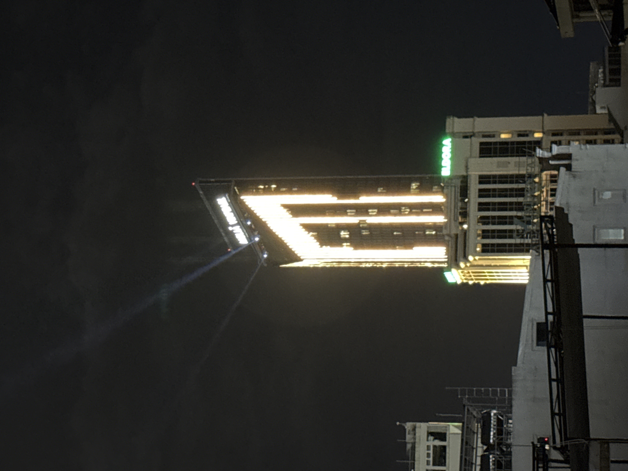 This media file depicts a nighttime scene in Thuận Hóa Ward, Vietnam. The primary focus is a large, illuminated building facade displaying a prominent, abstract light installation that resembles a stylized letter "E." This installation is comprised of bright yellow and white lights, arranged in horizontal and vertical segments against the dark night sky.
Adjacent to this illuminated structure, another building is visible with a vertical green illuminated sign that reads "ELDORA." Other buildings with lit windows are scattered throughout the frame, suggesting an urban environment. Beams of light, possibly from spotlights, emanate from the lower left of the frame, directed upwards towards the illuminated facade, creating a dramatic visual effect. The overall impression is one of an architectural light show or advertisement at night. The sky is dark and appears clear, indicating a dry night.