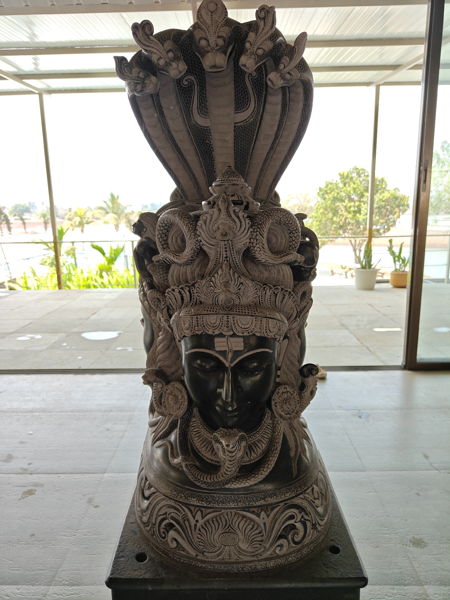 The image shows a carved stone statue of a deity with multiple snake heads, located indoors in Kalote Mokashi, India. The statue features a dark-colored human-like face with closed eyes and a forehead marking, adorned with a crown or headdress. Above the head, multiple cobra snakes are depicted with raised hoods. The statue rests on a dark-colored base with carved detailing. The background shows a bright, outdoor scene through a large window, revealing some greenery and buildings. The foreground features a tiled floor and a window frame. The statue is well-lit, emphasizing the intricate carvings and details.