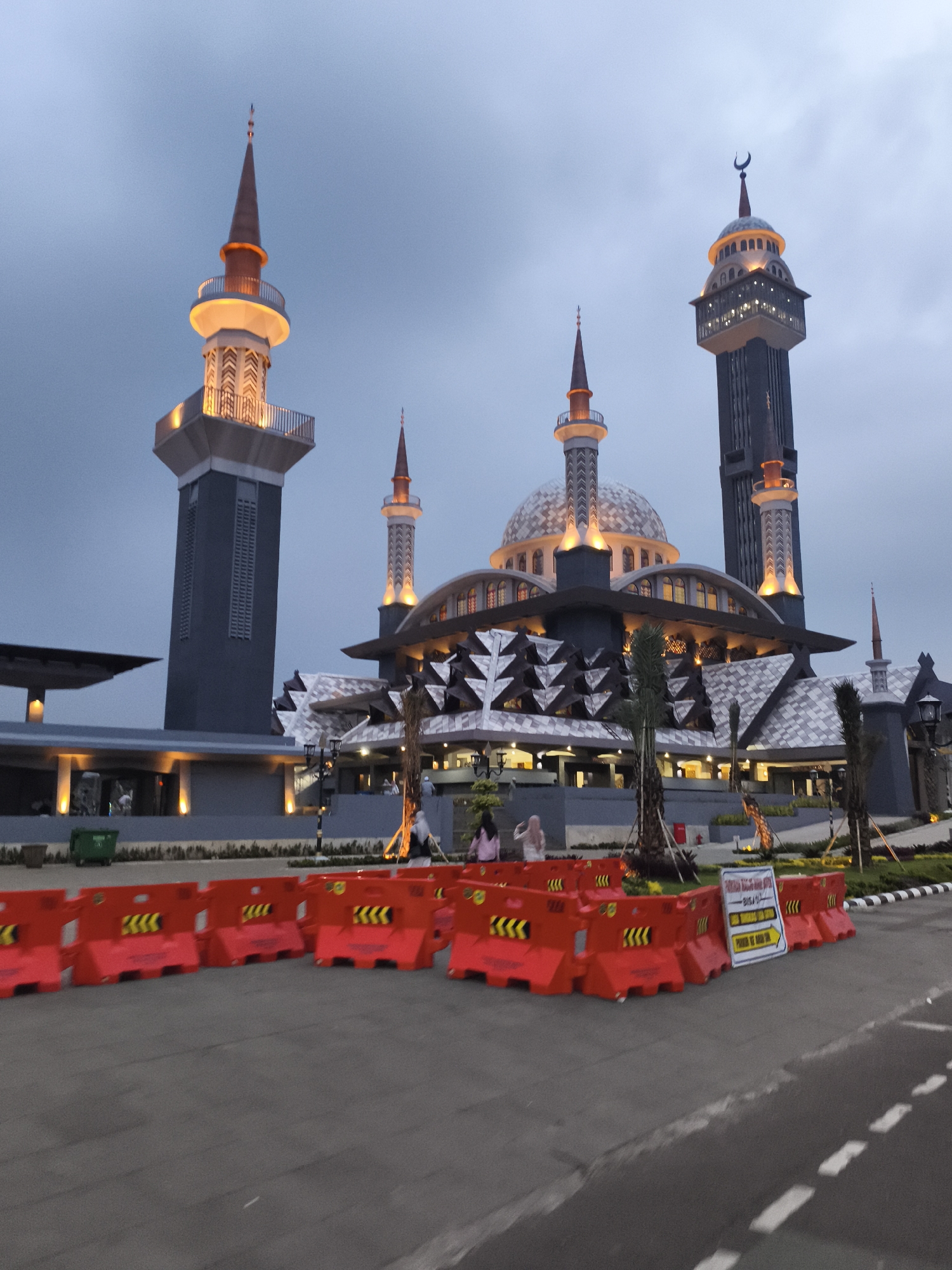This image captures a grand, modern mosque complex in Cibinong, Indonesia, illuminated at dusk or early evening. The sky is a uniform grey-blue, suggesting an overcast day transitioning into night, with the mosque's warm artificial lights prominently on.
The main subject is the mosque itself, featuring a large central dome surrounded by several smaller domes and multiple distinct minarets. The architecture blends traditional Islamic elements with contemporary design, including intricate geometric patterns on the building's facade. Two tall minarets stand out: one on the far left with a tapering spire and an illuminated circular balcony, and another on the far right which is a very tall, dark grey structure with an elaborate, brightly lit upper section crowned with a crescent moon finial. Other smaller minarets are integrated into the main structure, also brightly lit.
In the foreground, a line of bright orange plastic road barriers, marked with yellow chevron patterns, stretches across the width of the image, separating the viewer's position from the mosque grounds. Attached to one of these barriers is a white sign with black text that reads "BISA PARKIR DI AREA INI" (Can Park In This Area) and "PARKIR DEKAT MASJID" (Parking Near Mosque), accompanied by a right-pointing arrow, indicating a nearby parking facility.
Several people are visible in the middle ground, near the mosque's entrance and on elevated walkways, appearing to be visitors or congregants. Two individuals, one in a pink shirt and another in a light-colored hijab, are clearly discernible on the left side of the main building. Green landscaping, including palm trees, surrounds the complex. A green trash bin is also visible near the base of the mosque complex on the left. The scene evokes a sense of grandeur and peaceful evening activity at a significant religious landmark.