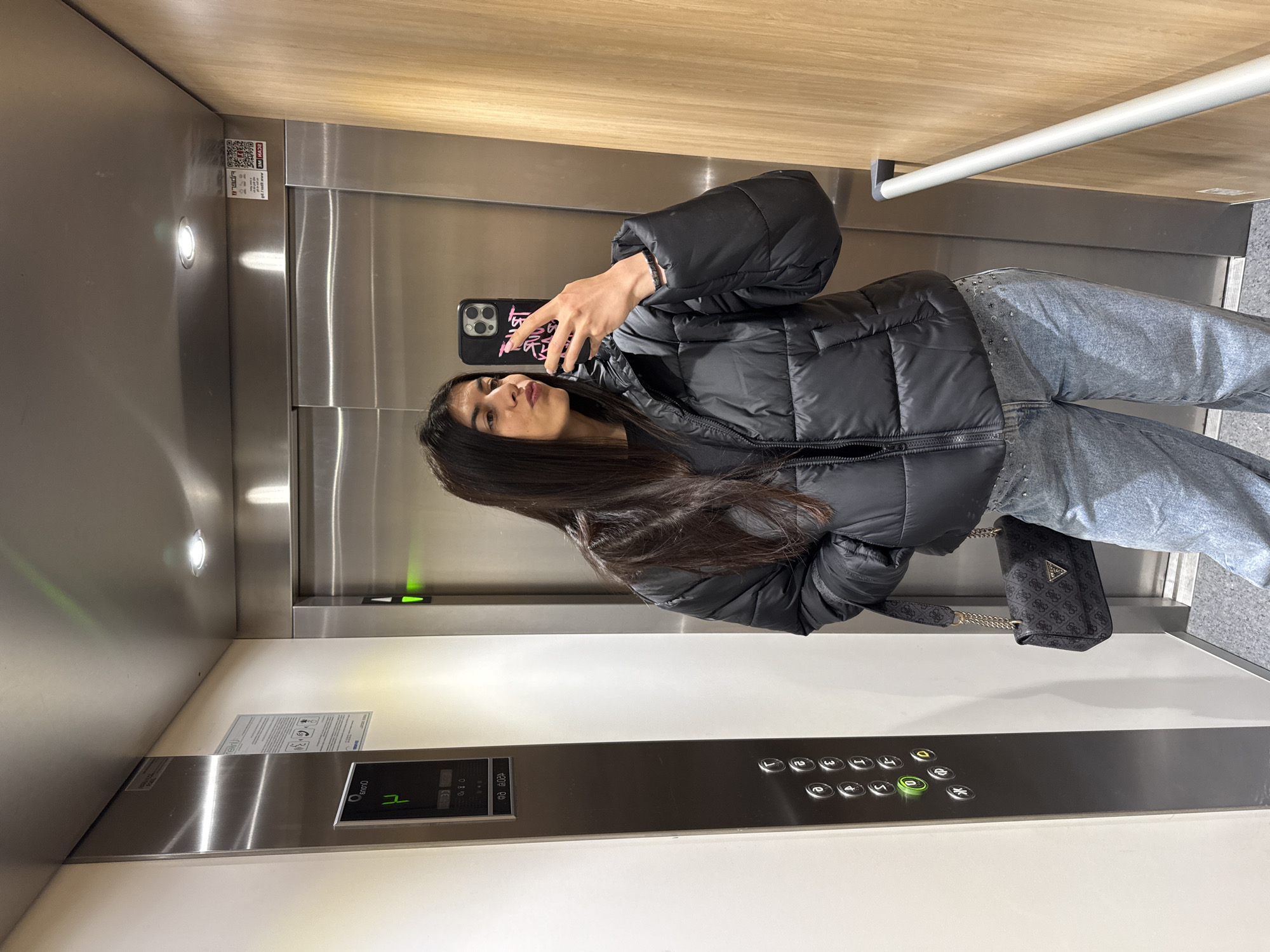 A woman takes a selfie in an elevator. She is wearing a black puffer jacket and light-wash jeans. She is holding a black smartphone with a pink design on the back. She has long, dark hair. A Guess handbag hangs from her shoulder. The elevator has a stainless steel interior and a panel of buttons. The number '1' is illuminated on the panel. A sticker with a QR code is visible on the upper-left side of the elevator. The location is unknown, there is no information that relates the location to Sofia, Bulgaria.