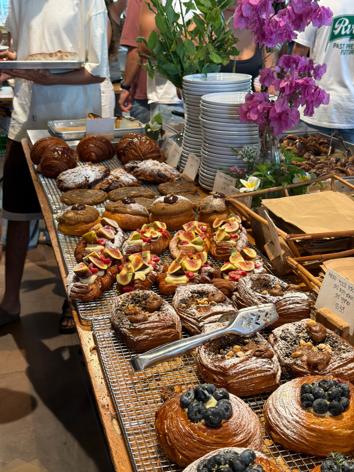 The image depicts a display of assorted pastries, likely at a bakery. In the foreground, a variety of danishes and croissants are arranged on metal cooling racks. Some pastries are topped with fruit like figs and raspberries, while others are dusted with powdered sugar and nuts. There are also some round pastries topped with blueberries. In the background, a stack of white plates is visible, as well as a vase of purple flowers and greenery. Partially visible to the left is a person holding a tray of bread, and to the right, another person is standing. The overall setting appears to be indoors, with a wooden counter and a concrete floor. The scene suggests a retail environment, possibly a bakery or cafe. The presence of Hebrew text on some labels implies the location is in Israel, potentially in Kfar Monash, given the prompt.
