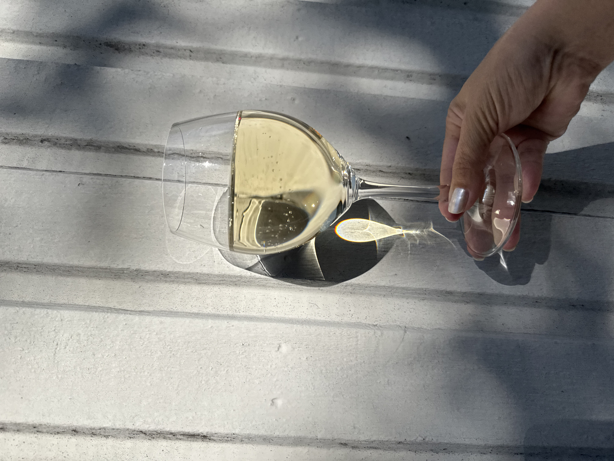 A close-up, eye-level shot shows a hand with pearl-white nail polish holding a stemmed wine glass filled with a pale yellow liquid, likely white wine or champagne. The glass is positioned in the center right of the frame, with the liquid catching the sunlight and reflecting a rainbow prism onto the textured, grey background. The background is a corrugated metal surface with vertical ridges, some of which are illuminated by the sun while others are in shadow. The shadows of the glass and its contents are cast onto the wall, creating a dramatic visual effect. The overall impression is one of a bright, sunny day. The location of Österskär, Sweden is not discernible from the image itself.