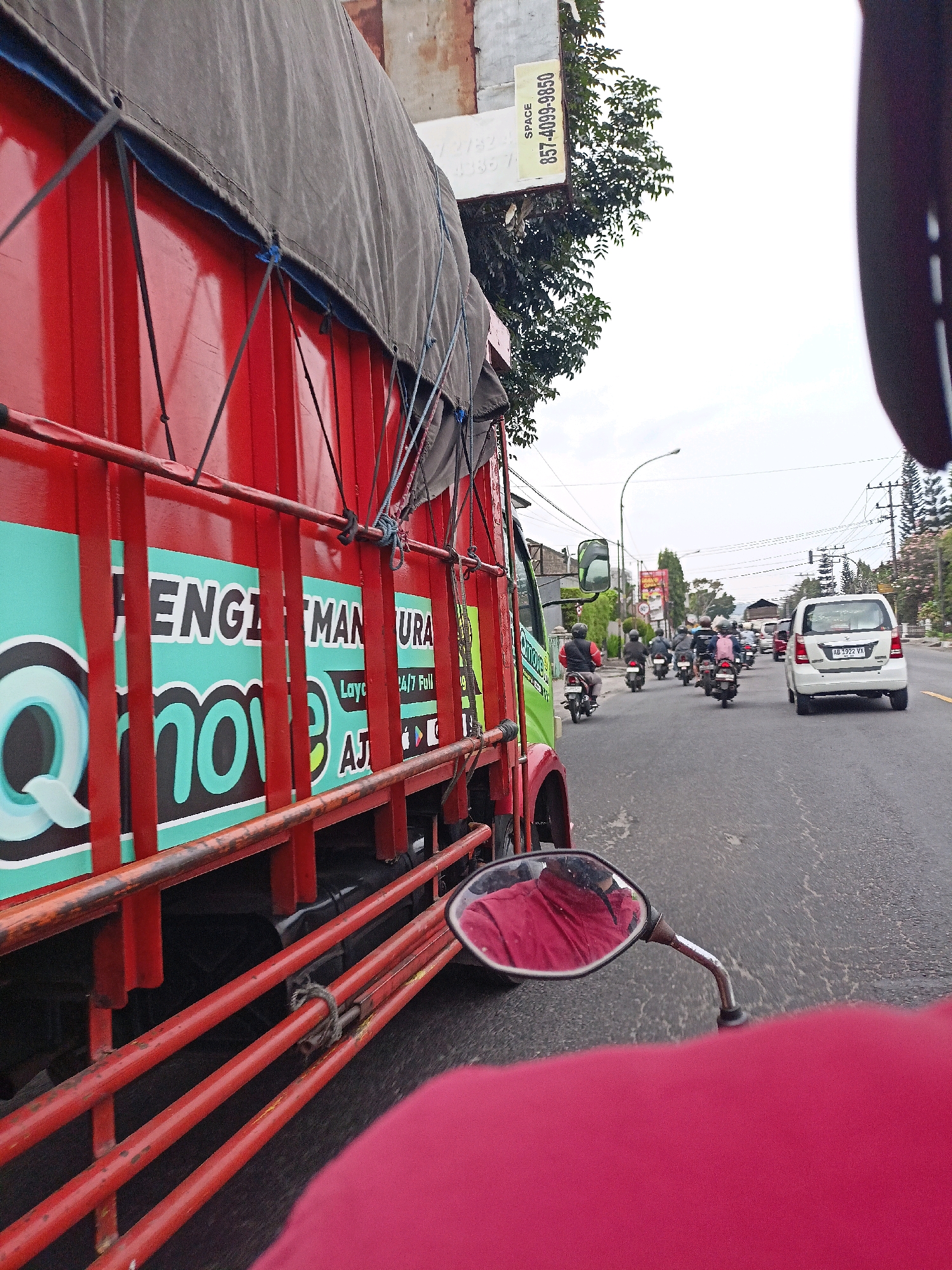 The image captures a street scene in Magelang, Indonesia. In the foreground, a red truck with a grey tarp covering its cargo is prominently featured on the left side. The truck has teal and white lettering on its side, which is partially visible and appears to be advertising a service called "Qmove" and mentions "LAYANAN 24/7 Full". A motorcycle rider, whose perspective is captured, is traveling behind the truck. The rider is wearing a red garment, and their reflection can be seen in the motorcycle's rearview mirror. Behind the truck, a line of motorcycles and a white car are traveling on the road. The environment appears to be urban, with buildings and trees visible in the background, along with overhead electrical wires and a street lamp. The sky is overcast. The license plate on the white car reads "AB 1922 VX". A yellow sign with black text, including "SPACE 357-4099-9650", is attached to a pole on the right side of the truck.