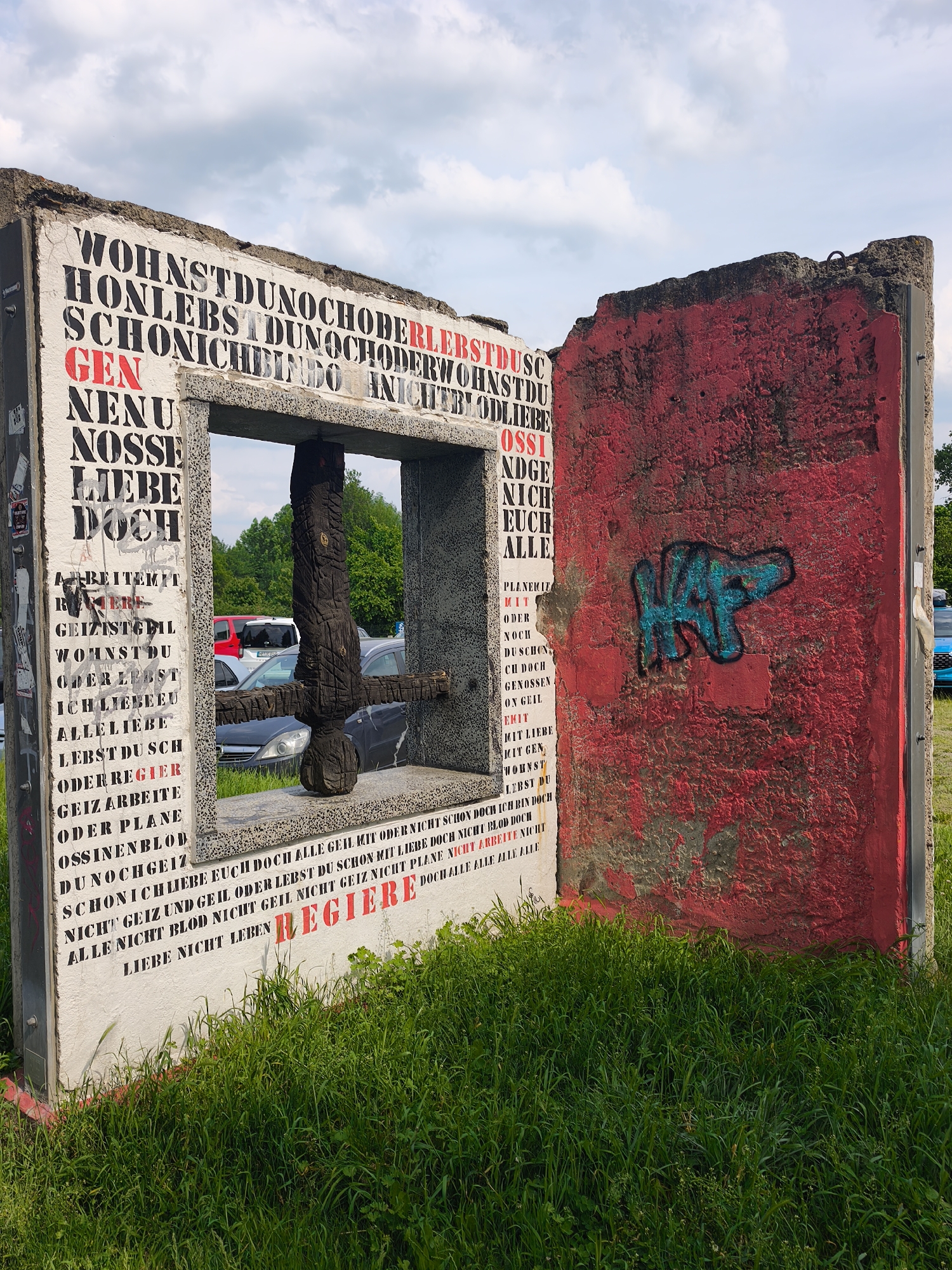 Here is a detailed description of the image:
The image captures a weathered, graffiti-covered art installation set against a backdrop of a parking lot and greenery, under a bright sky. The installation appears to be composed of concrete blocks, possibly remnants of a structure, with one block painted red and another white. The white block is densely covered in black and red text, possibly lyrics or quotes, in German. There is a wooden sculpture, resembling a cross or a symbolic sword, positioned within the opening between the blocks, with cars and trees visible through it. The red block features an abstract graffiti tag in blue. The installation is set on grass. The focus is on the contrast between the worn structure and the vibrant text, as well as the juxtaposition of the sculpture with the modern cars visible in the background.