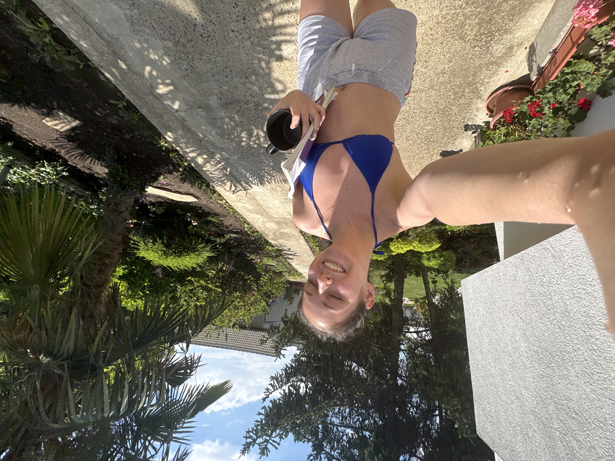 A young woman is captured in a selfie, her face turned towards the camera with her eyes closed and a wide smile. She is wearing a vibrant blue bikini top and light gray shorts. In her right hand, she holds a black mug and a stack of papers. The setting appears to be an outdoor patio or walkway, with lush greenery and trees surrounding her. Sunlight casts dappled shadows on the concrete surface. In the background, a portion of a house with a tiled roof is visible, along with a clear blue sky dotted with clouds. The scene evokes a sense of relaxation and enjoyment on a warm, sunny day. Based on external information, this location is in Bulgarovo, Bulgaria.