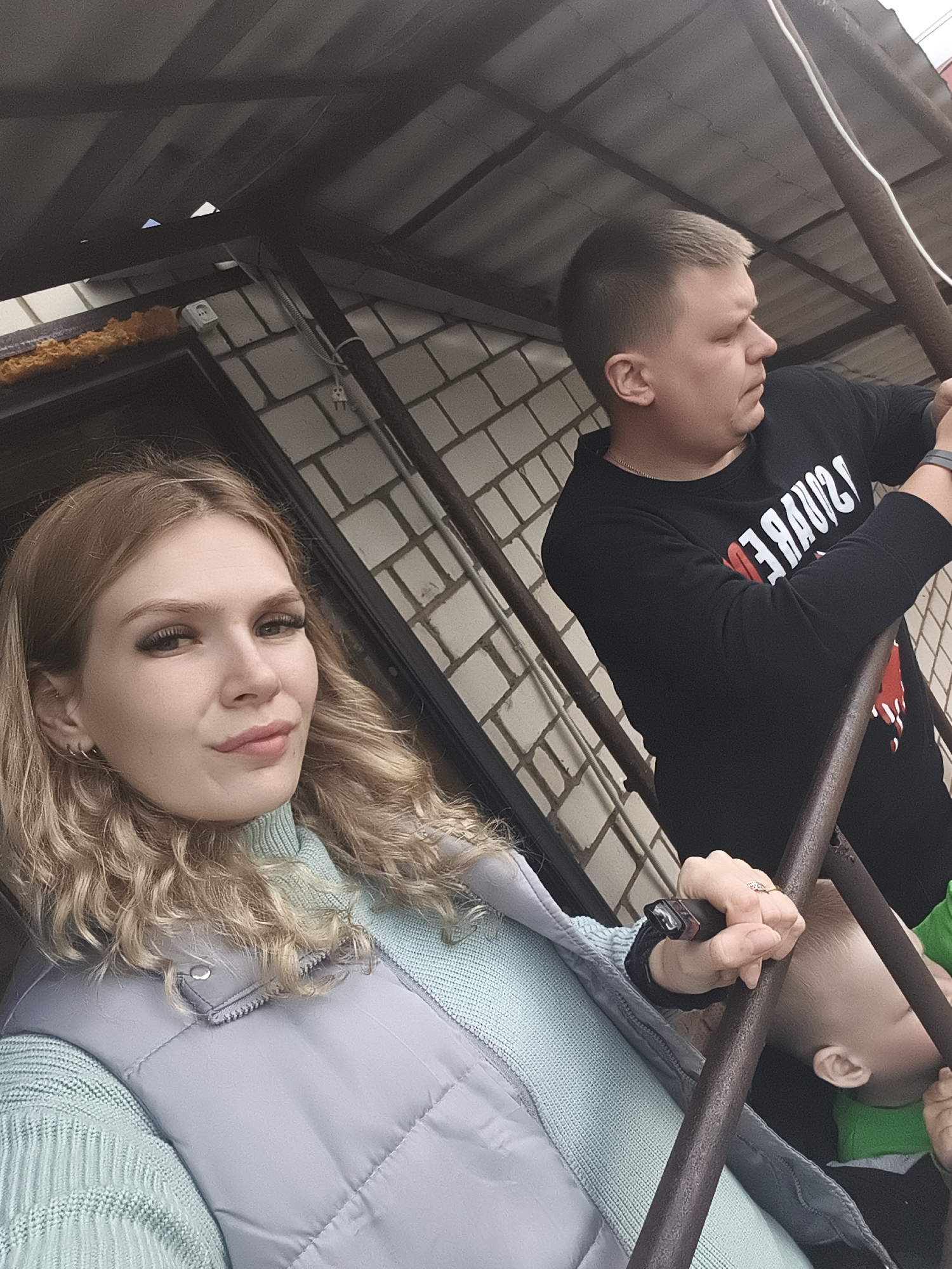 The image captures three individuals—a woman, a man, and a young child—in an outdoor setting, likely on a staircase or platform with metal railings, in Русский Вожой, Russia. The lighting suggests daytime conditions, possibly overcast or shady.
In the foreground, slightly to the left, a woman in her 20s or early 30s is visible. She has long, curly blonde hair and is wearing a light mint green turtleneck sweater under a light grey puffer vest. She is looking directly at the camera with a relaxed expression, her face made up with eyeliner and lip gloss. She wears small hoop earrings and a ring on her right ring finger, and holds a dark object in her right hand.
Behind her and to the right stands a man, likely in his 30s, with short, light brown hair. He is wearing a black long-sleeved sweatshirt with white, mirrored text on the front, which appears to be a reversed word, possibly "SQUARE" or similar, with some red detailing below. He is looking off to his right (viewer's right). A watch or fitness tracker is visible on his left wrist.
Partially visible at the bottom right, below the man, is the top of a small child's head and ear, wearing a green garment.
The background features a white brick wall and a dark, corrugated roof structure or canopy overhead. An electrical outlet with a white cable and plug is visible on the brick wall, along with some orange foam insulation. Rusty metal railings divide the foreground from the background, suggesting they are on an elevated structure. The overall scene appears to be a casual moment captured in a functional, possibly utilitarian, outdoor area.