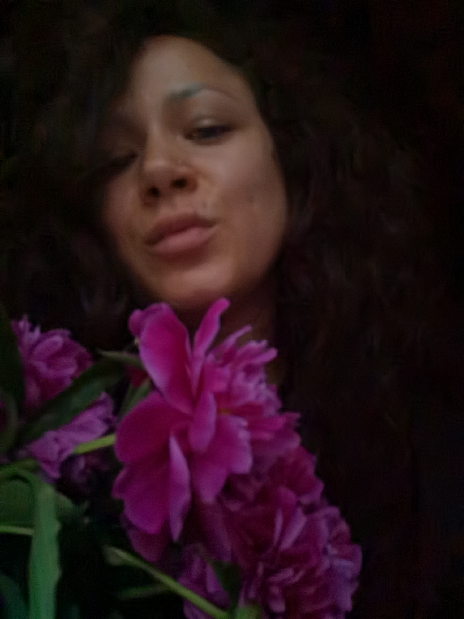 A close-up, dimly lit, and slightly blurred indoor portrait of a woman holding a bouquet of pink peonies. The woman has dark, curly hair and is looking towards the camera with her lips pursed. The peonies are vibrant and take up a significant portion of the foreground. The background is dark and indistinct, suggesting an indoor setting with low lighting. There is no discernible indication of Chaikovsky, Russia, in the image.