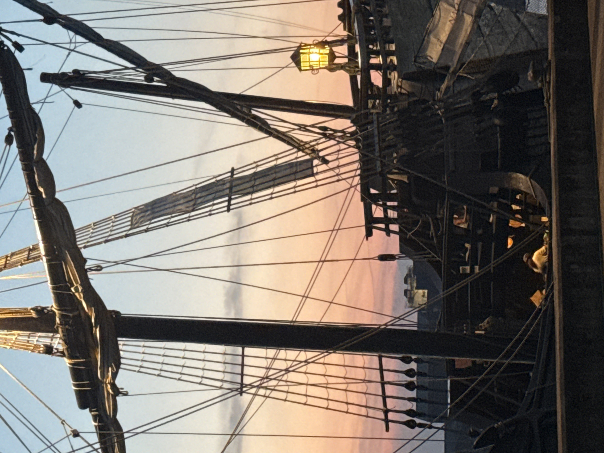 The image shows a tall ship docked at sunset in Great Yarmouth, United Kingdom. The ship is dark and detailed with many ropes and masts. A yellow lantern is lit on the ship. The sky is a gradient of orange and blue. There are buildings in the background, suggesting a harbor or waterfront area. Two people are visible on the deck of the ship, one wearing a yellow shirt.