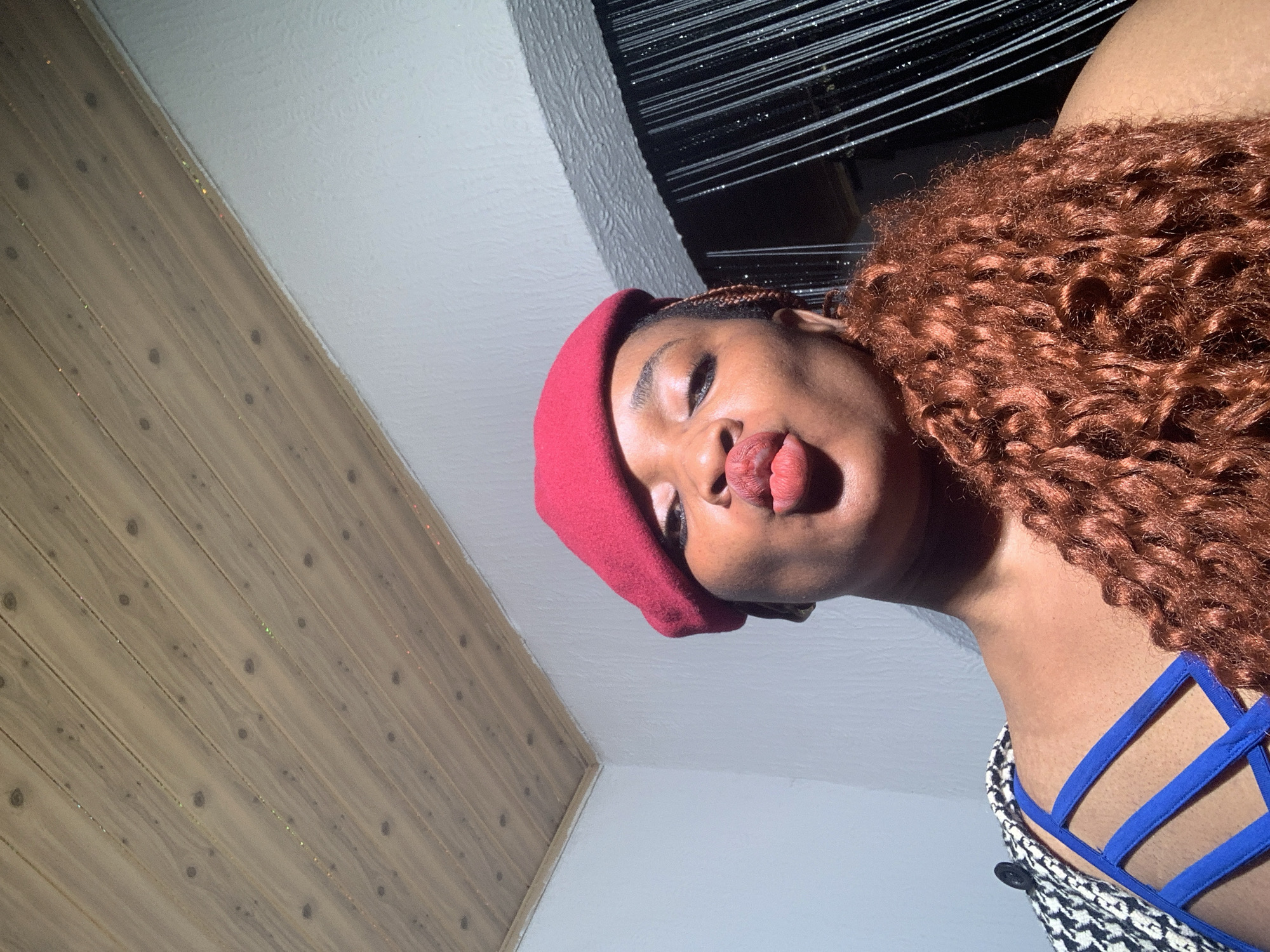 This low-angle, tilted portrait captures a young individual, likely female, with dark skin, posing playfully with her tongue extended slightly. She is seen wearing a vibrant red beret and features voluminous, tightly curled reddish-brown hair. Her clothing includes distinct blue straps of a top worn over a black and white patterned garment. The subject's face is illuminated by bright lighting, suggesting a daytime setting, which casts gentle shadows and highlights her features and hair.
The background of the indoor scene is split diagonally. To the left, there's a wall or ceiling section composed of light brown wooden planks with visible knots and grains. To the right, a plain, lighter-colored (possibly white or light grey) wall or ceiling surface is present. In the upper right background, a dark, horizontally textured element, possibly window blinds or a decorative feature, adds depth. The overall mood conveyed by the subject's expression is playful and confident. There is no visible text in the image. The photo is situated in Barakin Akawo, Nigeria.
