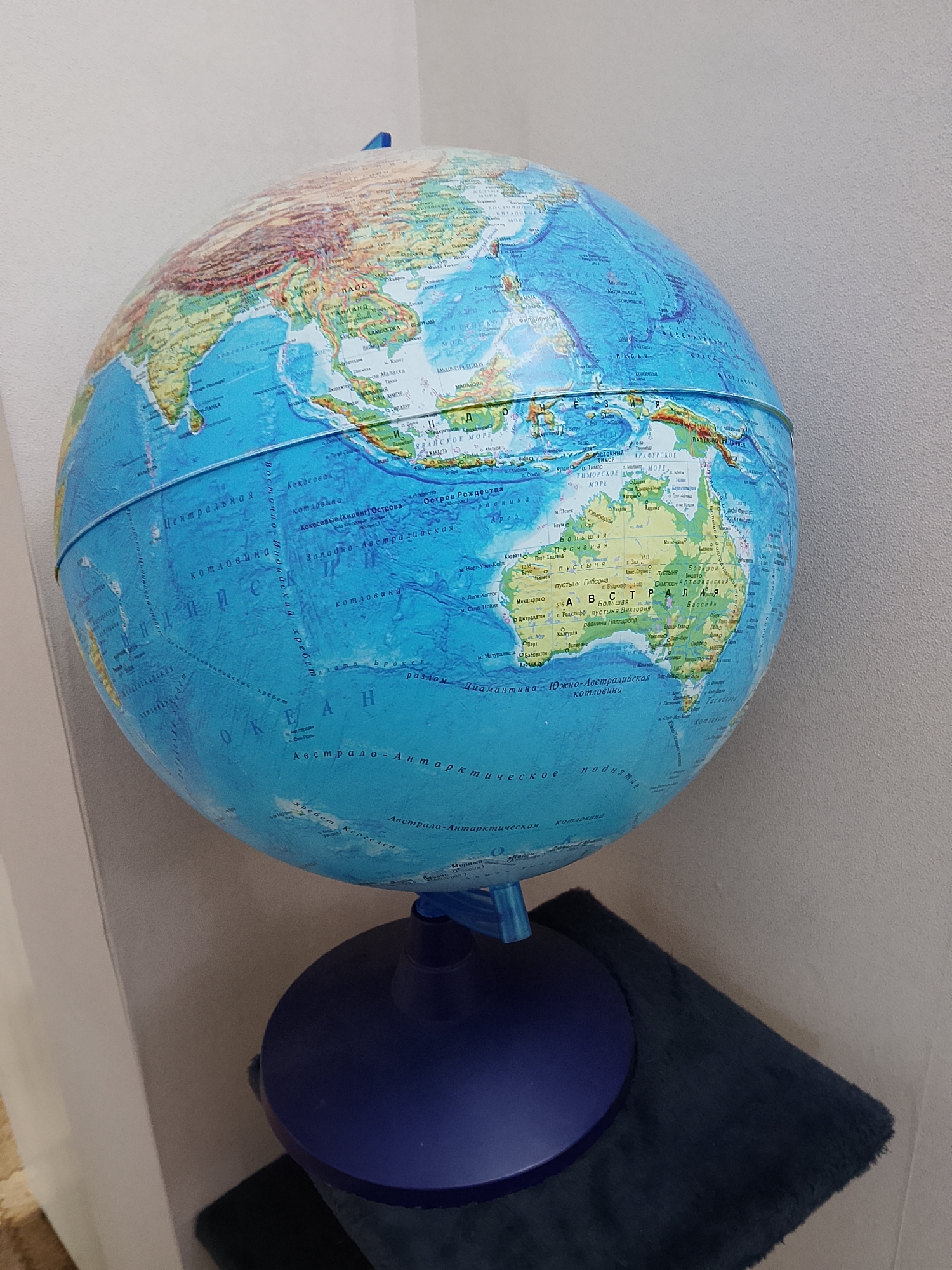 This media file features a globe of Earth, predominantly showcasing Southeast Asia and Australia. The globe is presented in a slightly tilted, upward-facing angle. It is mounted on a dark blue stand, which itself rests on a dark navy blue, fuzzy mat. The background consists of a textured, pale grey wall, and to the left, a hint of a patterned carpeted floor.
The visible text on the globe is in Russian and includes geographical names such as "ЛАНКА" (Lanka, likely referring to Sri Lanka), "МЬЯНМА" (Myanmar), "ТАЙЛАНД" (Thailand), "КАМБОДЖА" (Cambodia), "Океан" (Ocean), "Австралия" (Australia), and various sea and ocean names like "ЯВАЙСКОЕ MOPE" (Java Sea), "ТИМОРСКОЕ MOPE" (Timor Sea), and "Австрало-Антарктическое поднятие" (Australasian Antarctic Rise). City names like "Перт" (Perth) and "Алис-Спринге" (Alice Springs) are also legible.
There are no people or animals visible in the image. The lighting appears to be from an artificial source, creating a slightly diffused light. The overall setting suggests an indoor environment, possibly a home or an educational space. The focus is entirely on the globe and its geographical representation.