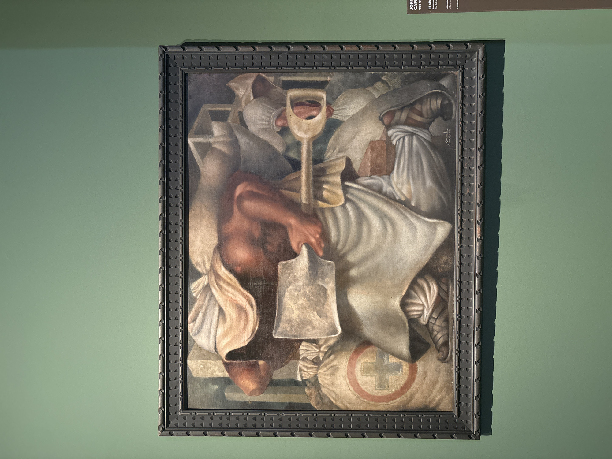 The image shows a painting in a museum. The painting is framed and hangs on a green wall. The painting depicts a muscular, dark-skinned figure draped in white cloth, carrying a large shovel and a sack with a red cross symbol. The figure appears to be in a construction or labor setting, with bricks and other materials visible in the background. The style of the painting suggests a mural or a piece from a larger work. A label to the right of the painting indicates it is titled "The Bricklayer" by Jorge Camarena, and its dates are 1908-1975. The label also mentions "El muralista de la revolucion social," indicating the artist's association with social revolution. The artwork is located in Rome, Italy, specifically at the Galleria Nazionale d'Arte Moderna e Contemporanea.