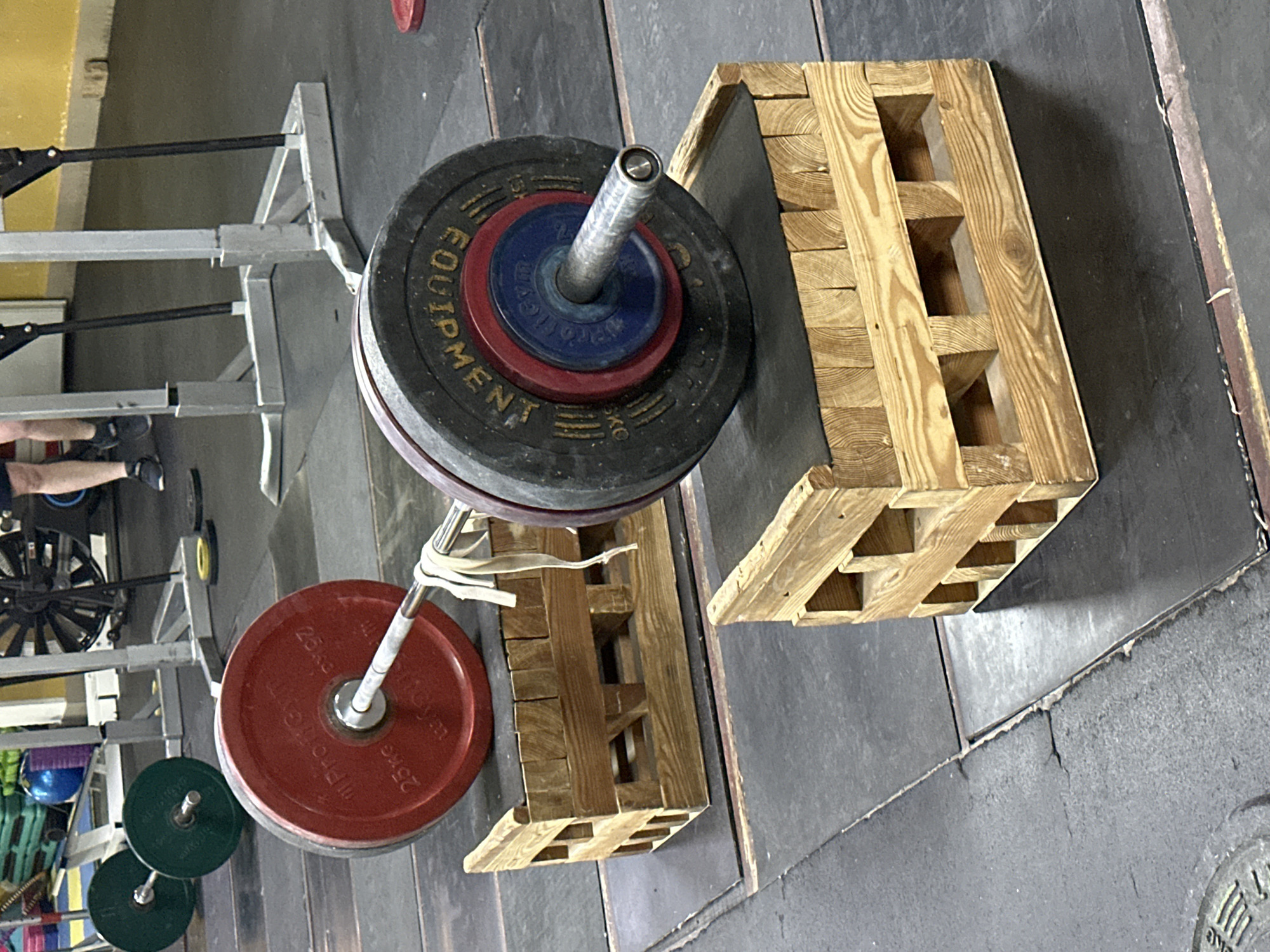 The image depicts a weightlifting setup in a gym. A barbell loaded with several weight plates of varying sizes and colors rests on wooden blocks. The weights range from black plates with "EQUIPMENT" printed on them, red plates marked "25 kg," and smaller green plates. The blocks are constructed of light-colored wood with square openings. In the background, a weight rack and a leg press machine are partially visible. The floor is made of dark-colored, textured rubber tiles. The scene is in a gym, but there is no information available in the picture to connect it to Blagoveshchensk, Russia.
