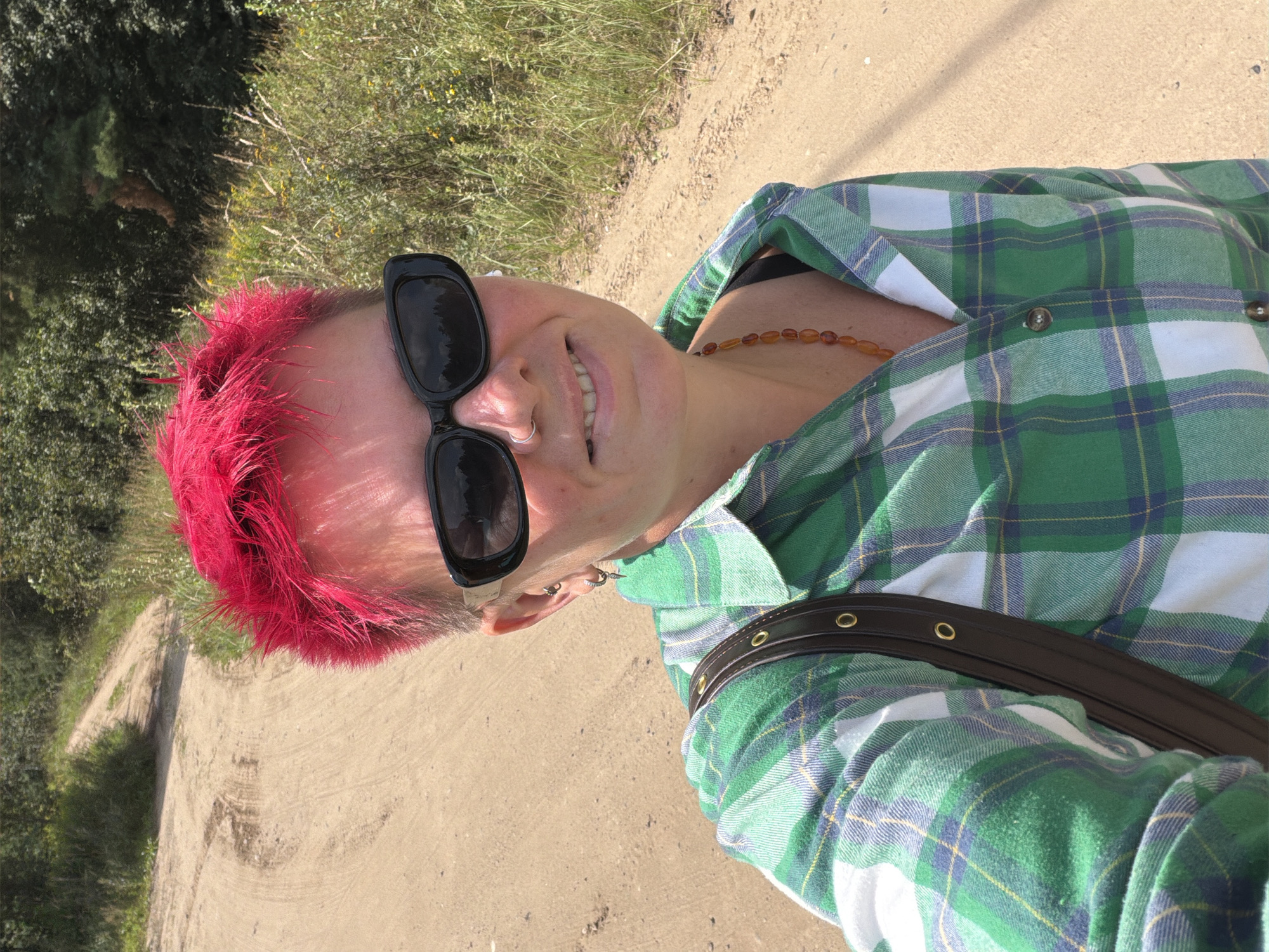 A woman with bright pink, spiky hair, wearing black sunglasses, a green and white plaid shirt, and an amber beaded necklace, takes a selfie outdoors. She has a silver nose ring and earrings. A dark brown strap of a bag is visible across her shoulder. The background shows a dirt road winding through tall grass and trees. The image appears to be taken in Chaikovsky, Russia, given the rural, natural setting. The lighting suggests it is daytime with the sun shining, casting shadows.