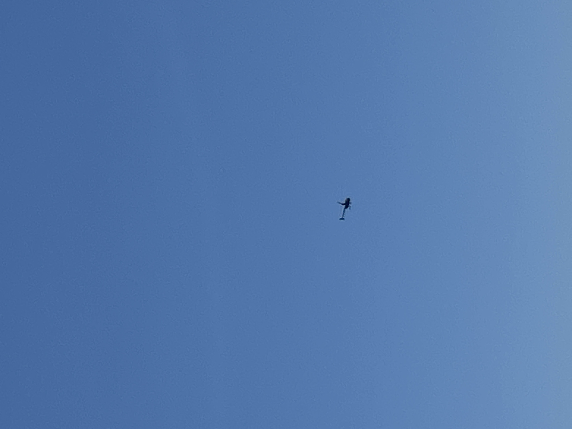 A small helicopter is captured in mid-flight against a clear blue sky. The helicopter is dark in color and appears to be a single-rotor model. It is positioned in the lower left quadrant of the image. The vast expanse of the sky dominates the composition, with no other objects or structures visible. The image was taken in Barcelona, Spain.