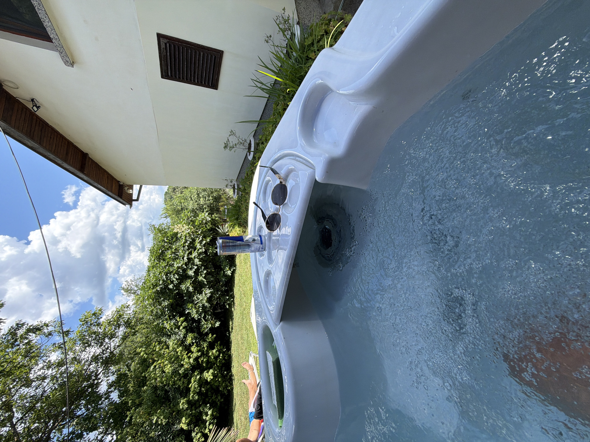 In Chavei, Bulgaria, a white, multi-level hot tub is filled with bubbling water. On the top level of the tub, a red bull energy drink can and a pair of sunglasses rest on the edge. In the background, a person lies on a lounge chair on a manicured lawn, surrounded by lush green trees and a white building with brown shutters. The sky above is bright blue with scattered white clouds.