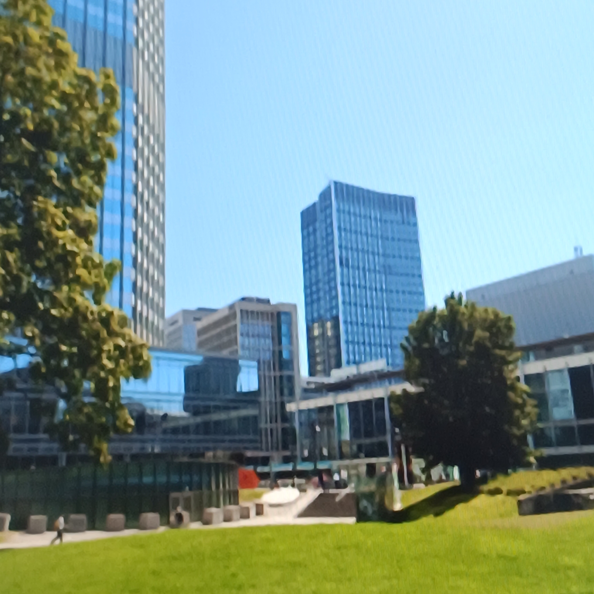 The image shows a bright, sunny day in a modern urban environment. In the foreground, a lush green lawn is visible with a pathway running through it. Along the pathway, several large, cube-shaped concrete planters are arranged. A lone figure, appearing to be walking, is on the pathway. In the midground, there are several contemporary buildings with reflective glass facades, indicating an office or commercial district. One prominent building is a tall, slender skyscraper with a blue-tinted glass exterior. To the right, a mature tree with dark green foliage casts a shadow on the grass. In the background, more of the cityscape unfolds with additional modern architecture under a clear blue sky. The overall impression is one of a clean, well-maintained urban park area adjacent to business buildings.