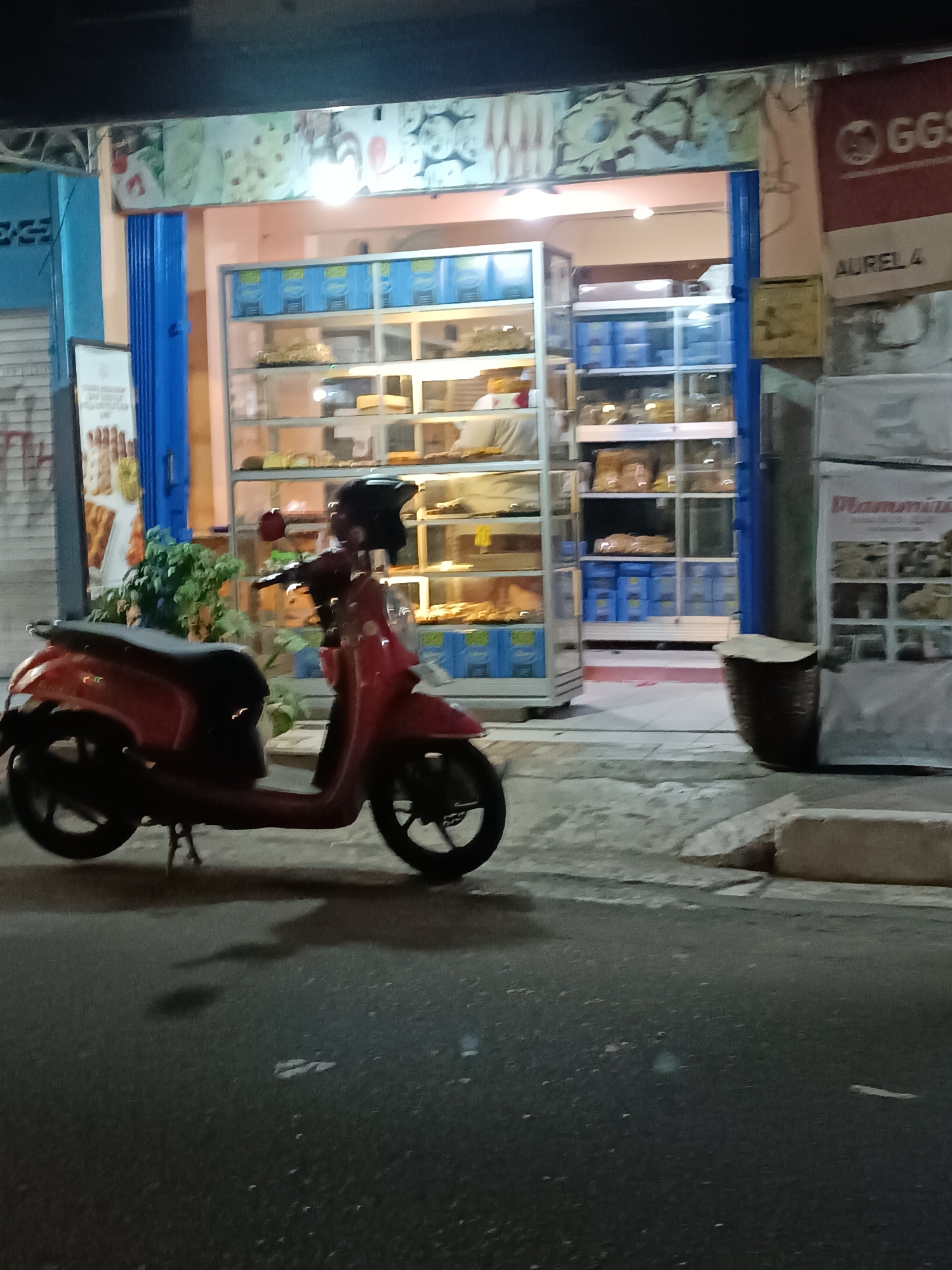 A red scooter is parked on the side of a street in front of a bakery in the city of Yogyakarta, Indonesia. The bakery has large glass display cases filled with baked goods. Above the entrance, there is a colorful advertisement for pastries. A sign on the right side of the building reads "GGS" and "AUREL 4". Another sign on the far right displays "Mammini". The street is paved with asphalt, and there is a concrete curb in the foreground. The scene is illuminated by artificial light, suggesting it is nighttime or dusk.
