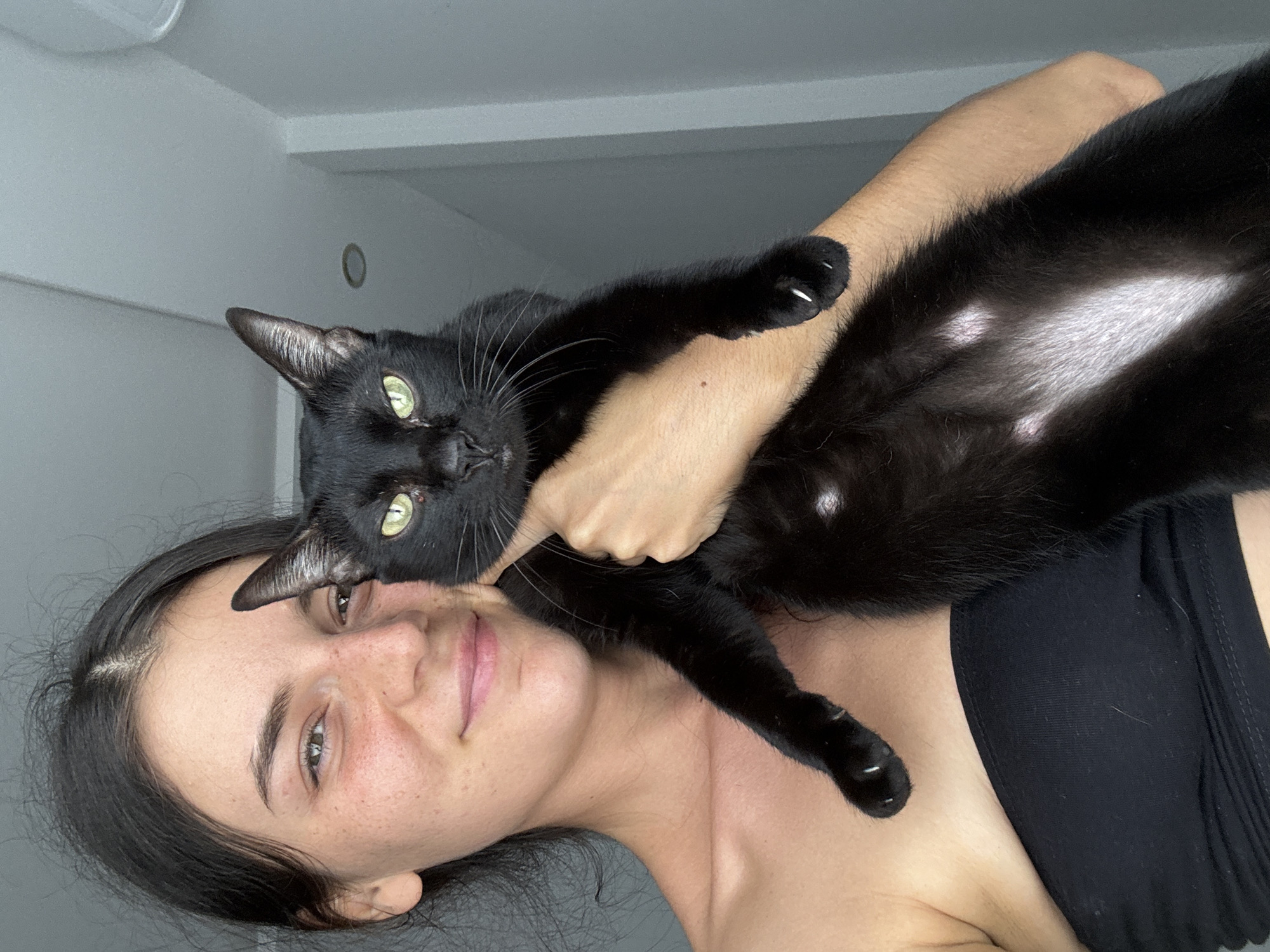 A young woman with dark hair and freckles is holding a black cat in her arms. She is wearing a black bandeau top and smiling at the camera. The cat has bright green eyes and white patches on its chest and belly. The background appears to be an indoor setting with a gray wall and some lighting fixtures. There is no indication of the image being taken in Mũi Né, Vietnam.