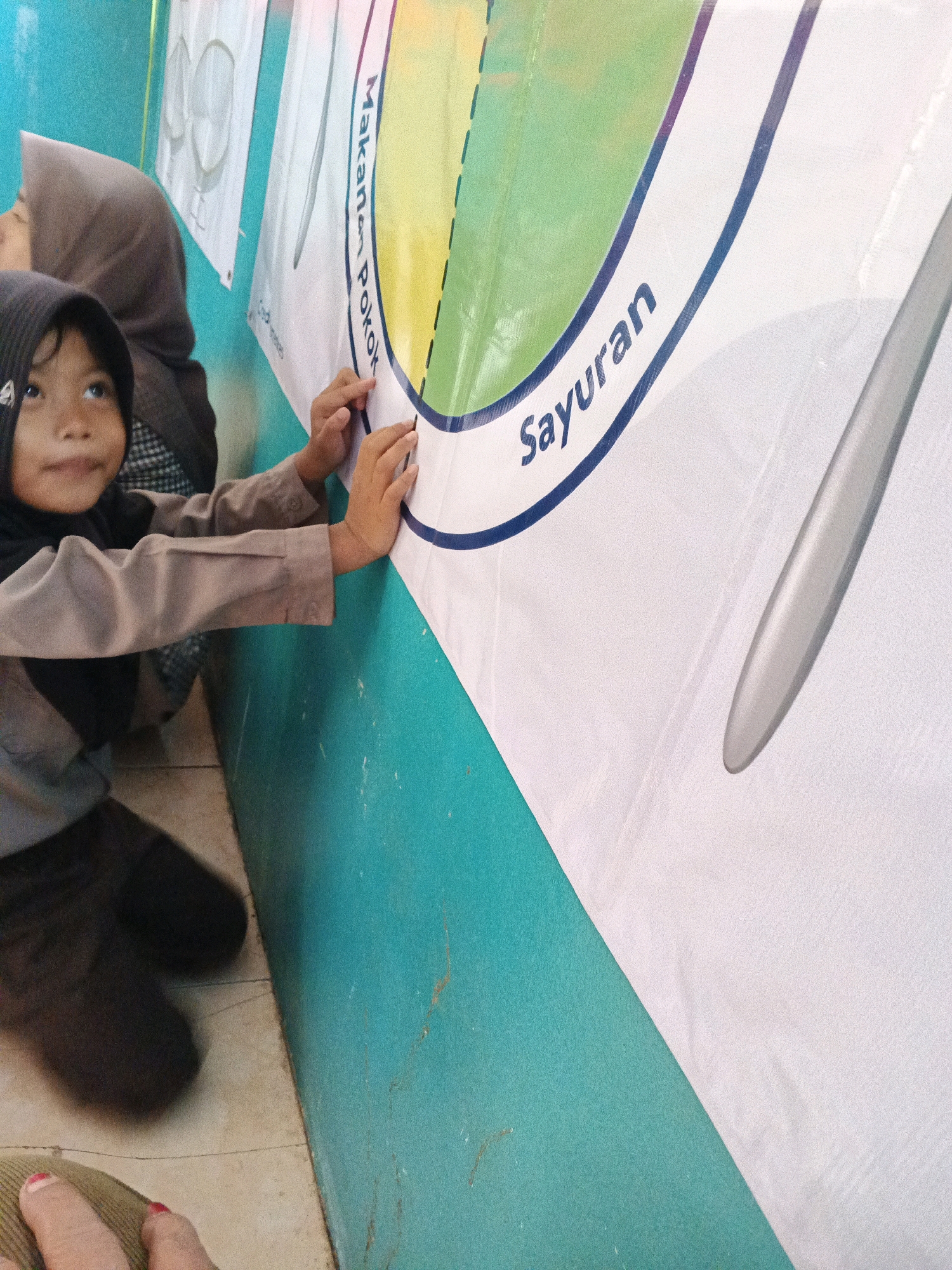 This image shows two young girls indoors, likely in a school setting in Warungkiara, Indonesia. The girl in the foreground, wearing a dark hijab and a light brown uniform, is looking to her left with her mouth slightly open. She is touching a large white banner with colorful circular designs and text. To her left and slightly behind her, another girl wearing a brown hijab is visible. The banner features the words "Makan" and "Sayuran" in Indonesian, indicating content related to food and vegetables. The walls are painted teal, and the floor appears to be tiled.