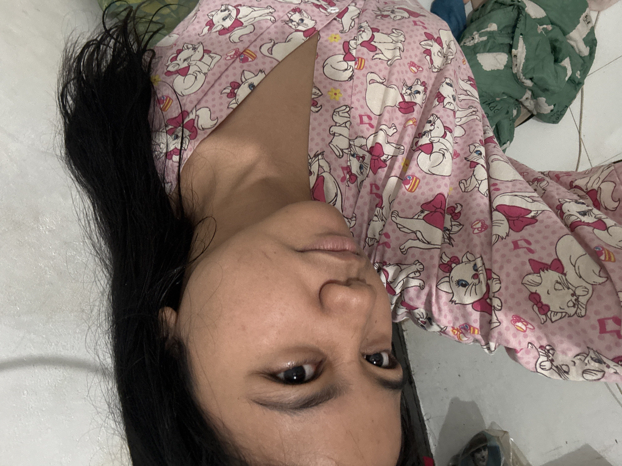 In North Jakarta, Indonesia, a medium shot captures a young woman from the chest up, looking directly at the camera. She is wearing a pink pajama top adorned with a pattern of white cartoon cats wearing pink bows. Her dark hair is down and falls over her right shoulder. The background is blurred, showing a white textured wall on the right and what appears to be a floor with some items on it to the left, including a green folded blanket with a white cloud pattern. The lighting is soft, and the overall scene suggests an indoor, casual setting.
