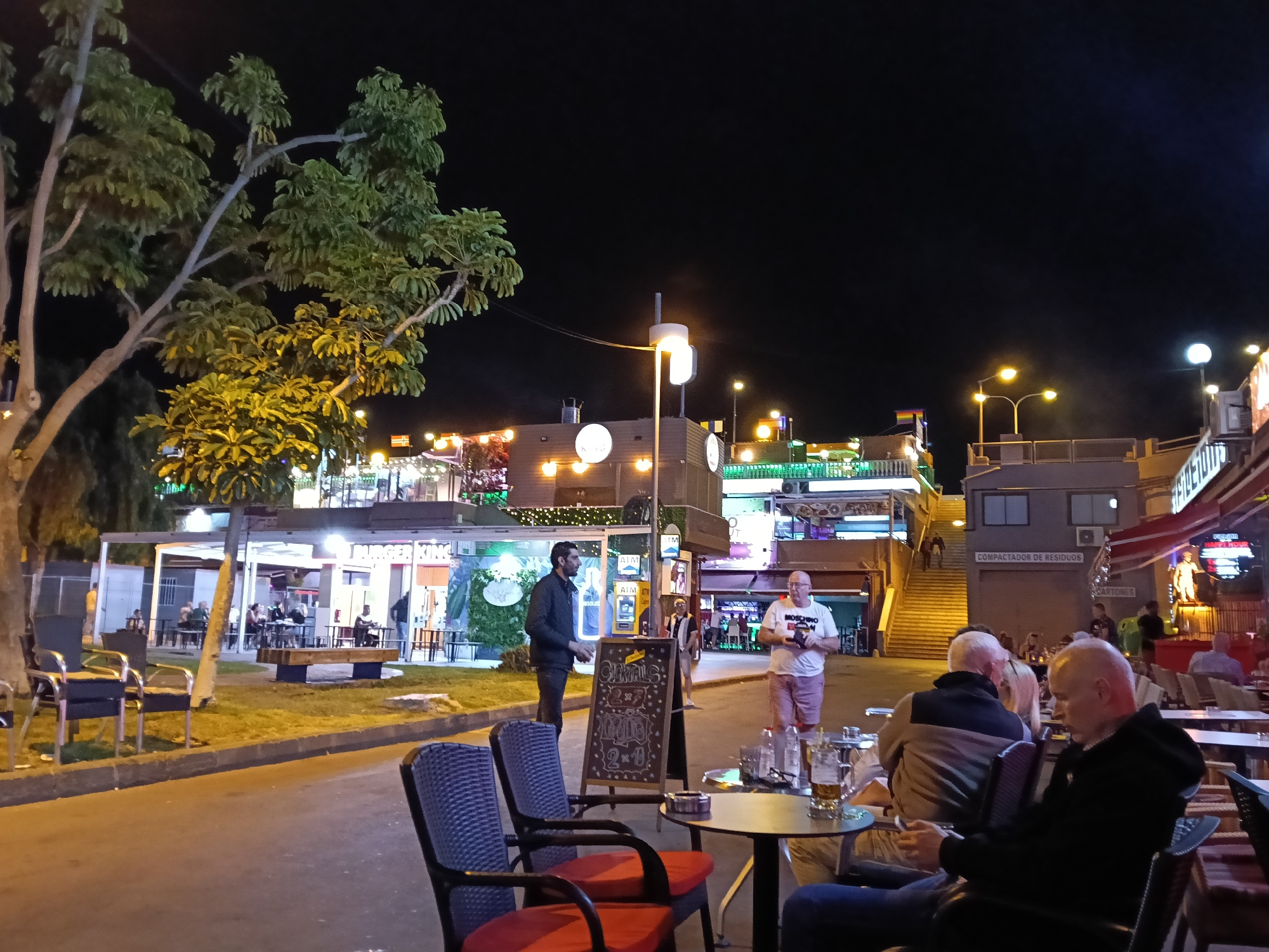 The image captures a vibrant night scene in an outdoor commercial area, likely in San Bartolomé de Tirajana, Spain. The setting is a bustling pedestrian zone with multiple levels of shops, bars, and restaurants, illuminated by numerous artificial lights.
In the foreground, several people are seated at outdoor tables, engaged in conversation and enjoying drinks, indicated by beer mugs and glasses on the tables. One man in the immediate foreground, with his back to the viewer, wears a dark hoodie and is looking down, possibly at a phone. Another man in a light brown jacket sits opposite him. To the right, another man, possibly holding a phone or camera, wears a white t-shirt with "MOSCHINO" printed on it and pink shorts.
Further back, a man in a dark jacket stands beside a blackboard A-frame sign that advertises "COCKTAILS 2x1". Beyond him, other individuals are seen walking or sitting at tables under sheltered areas. On the left, a large tree with green foliage is prominent, partially lit by the surrounding lights.
The background features multi-story buildings housing various establishments. A "BURGER KING" sign is clearly visible, along with multiple "ATM" signs. Bright streetlights and building facade lights cast a warm glow over the scene. A large set of stairs leads up to a higher level of the complex. On one building, text reads "COMPACTADOR DE RESIDUOS" (Waste compactor) and "CARTONES" (Cardboard boxes) below it. A rainbow flag is subtly visible atop a building in the mid-ground, suggesting an LGBTQ+-friendly establishment or area.
The atmosphere appears lively and relaxed, indicative of an evening out in a popular tourist destination. The weather seems mild and clear, suitable for outdoor dining and socializing.