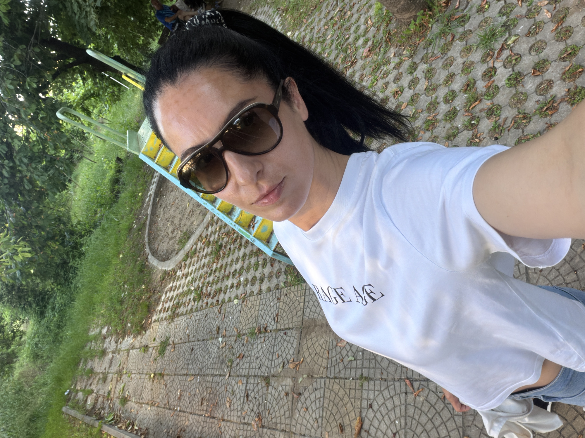 A female subject takes a selfie in an outdoor park or playground setting in Pleven, Bulgaria. She has dark hair styled in a ponytail and wears dark-lensed sunglasses with thick frames. Her attire consists of a white short-sleeved t-shirt with partial dark text (e.g., "RAGE AE") on the left chest and light blue denim jeans. Her gaze is directed towards the camera, which is held at an elevated angle, capturing her head and upper torso.
The ground surface comprises patterned interlocking concrete pavers and perforated concrete grass pavers, with grass and scattered dry leaves present in the interstices. A concrete curb borders a sandy/dirt area adjacent to the paving. In the background, a children's playground structure featuring faded yellow steps and light blue handrails is visible. Dense green foliage from multiple trees constitutes the background. Two partially obscured individuals are visible in the distant background among the trees; one wears a blue shirt. The scene is naturally lit during daytime.