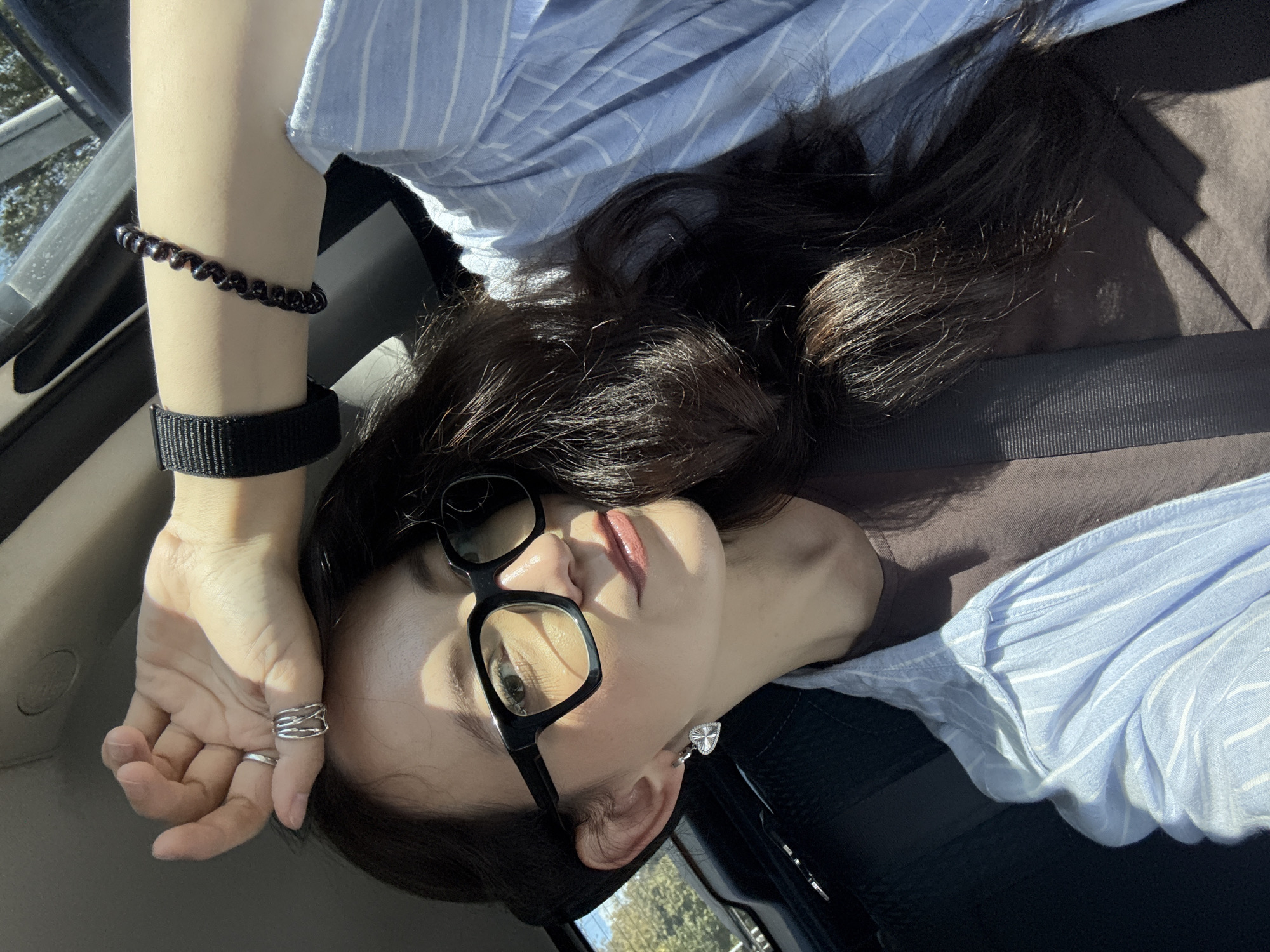 A woman is seen from the chest up, inside a vehicle, likely a car given the seatbelt visible. She has long, dark hair, wears glasses with thick black frames, and has on a light blue pinstripe shirt over a brown t-shirt. She has a heart-shaped earring in her left ear, a multi-ringed silver ring on her right ring finger, and a black beaded bracelet on her left wrist. Her right arm is raised and resting on her forehead, with her left arm bent, its elbow resting on the seat. The lighting suggests it is daytime, with sunlight casting shadows. The background is blurred but appears to be the interior of a car. There are no discernible landmarks or features that would place this image specifically in Kazan, Russia.