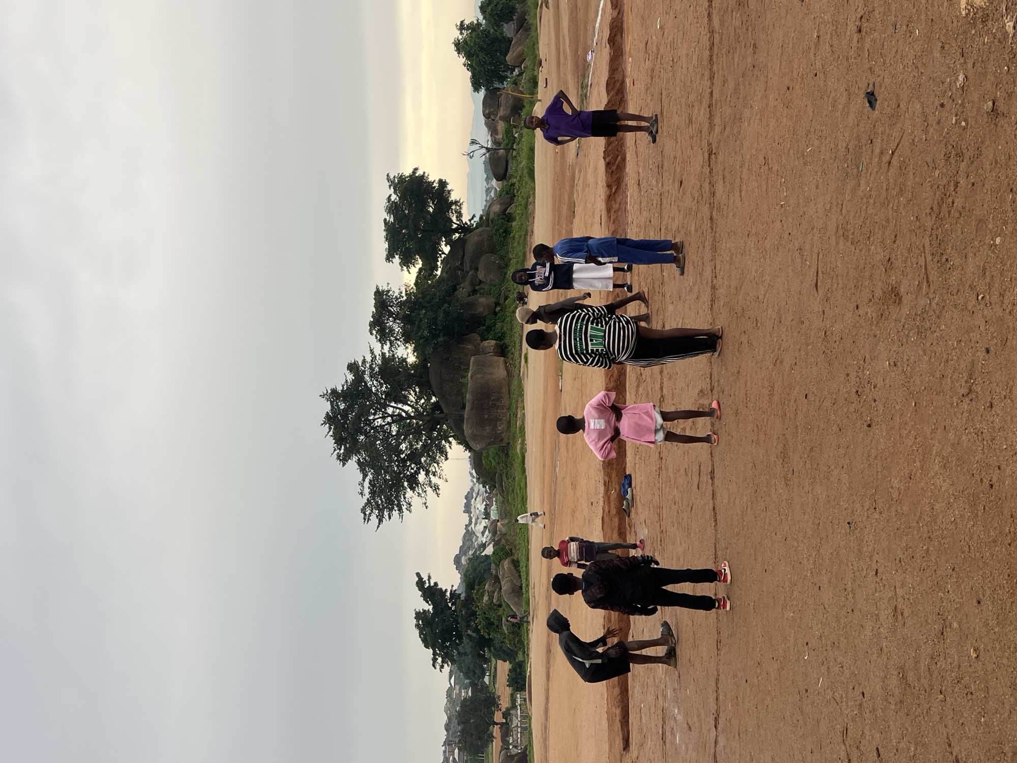 In Jos, Nigeria, a group of children are gathered on a dusty, reddish-brown field. The sky is overcast and pale. In the background, several large rock formations are visible, covered in sparse vegetation. Scattered trees grow on and around these rocks, with more dense foliage and distant buildings appearing further in the distance, suggesting a semi-urban or peri-urban setting. The children appear to be engaging in some form of activity, possibly a game, on the open ground. Some are standing and watching, while others are in various postures of movement or anticipation. Their clothing is casual, typical of children playing outdoors. The overall impression is of a community space where outdoor recreation takes place amidst the natural landscape of Jos.