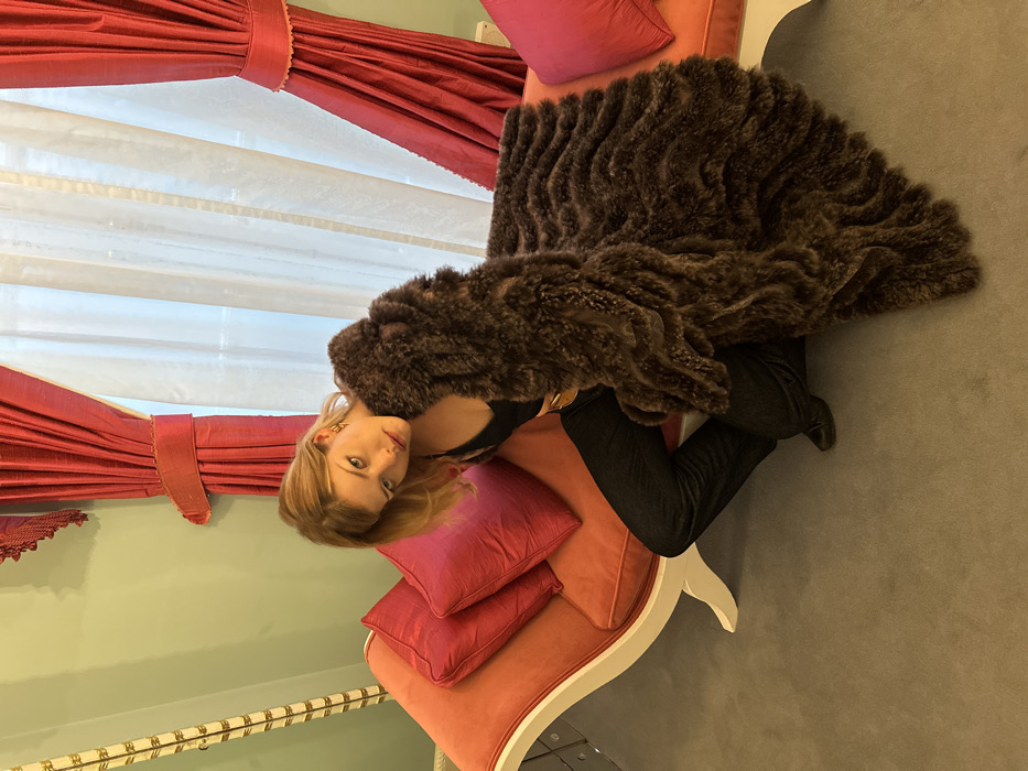 A woman sits on an ornate, pink and white chaise lounge, draped in a large, brown fur coat. She wears dark pants, a black top, and gold jewelry, with blonde hair framing her face as she looks toward the camera. Behind her, red drapes frame a sheer white curtain, adding to the room's decor. A light green wall is visible below the window, completing the scene's color palette.