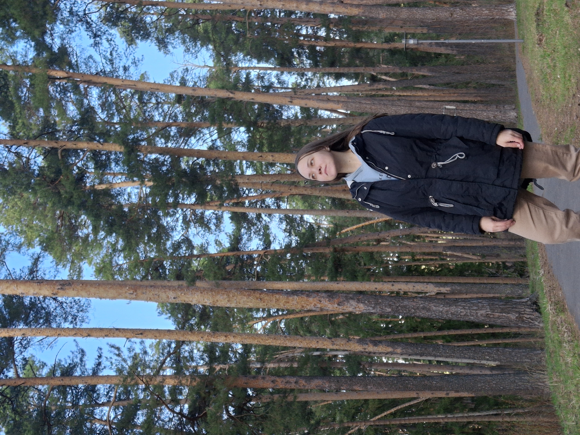 A young woman is standing on a path in a forest. She is wearing a black jacket, light blue shirt, and tan pants. The trees are tall and thin, with green needles. The sky is blue and visible through the branches. The path is made of asphalt and has grass growing on the edges. The scene is in Sarapul, Russia, as identified by the trees and other elements.