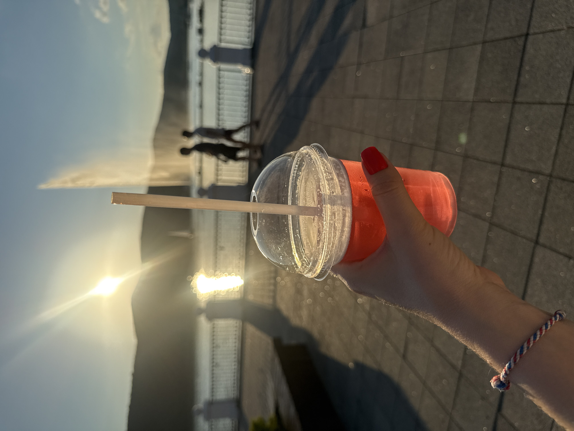 A person's hand, adorned with a red fingernail and a red, white, and blue braided bracelet, holds a clear plastic cup filled with a vibrant pink beverage. A paper straw is inserted into the cup. The background is softly blurred, suggesting a wide-angle shot.
The scene appears to be outdoors, near a body of water, with a waterfront promenade or walkway. In the distance, silhouettes of people can be seen walking. A fountain is spraying water into the air, creating a misty plume. The sun is low in the sky, casting a warm, golden light and creating lens flares. This indicates it is either sunrise or sunset. The ground is paved with square tiles, and long shadows are cast across it, further confirming the time of day.
The overall mood is serene and relaxed, possibly a leisurely stroll during golden hour. The location cues, combined with the context, suggest the picturesque waterfront of Abrau-Dyurso, Russia, a popular resort town known for its wine and scenic beauty. No visible text is present in the image.