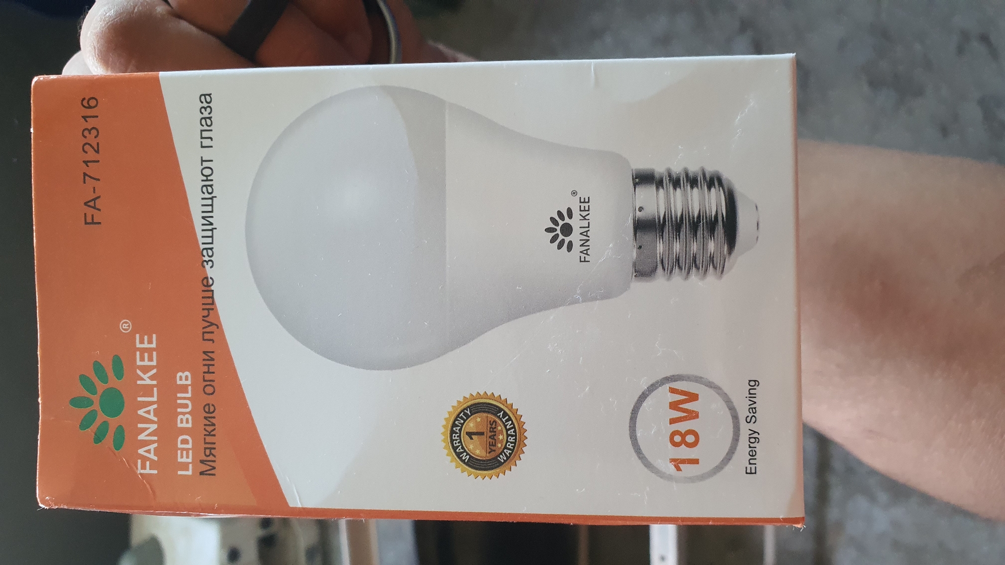 An orange and white box for a FAN ALKEE LED BULB is held by a person, whose hand and arm are visible on the right side of the image. The box is tilted, showing its front panel. Text on the box reads "FAN ALKEE", "LED BULB", "Мягкие огни лучше защищают глаза" (which translates to "Soft lights protect eyes better"), "FA-712316", a "1 YEAR WARRANTY" seal, "18W", and "Energy Saving". A white LED lightbulb is depicted on the box. The background is blurred and appears to be indoors.