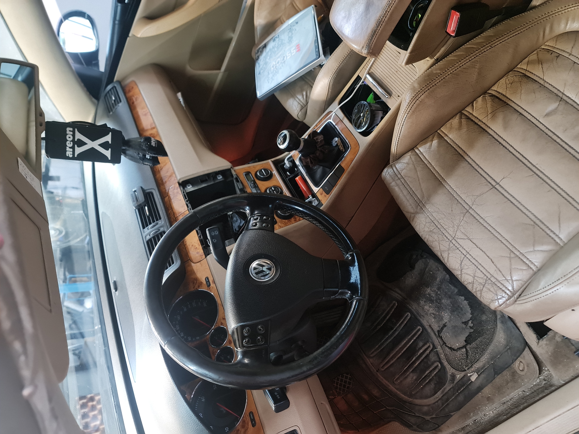 The image captures the interior of a car, likely a Volkswagen due to the emblem on the steering wheel, seen from a high angle looking towards the front passenger side. The setting appears to be a stationary vehicle during daytime, evidenced by the bright natural light filtering into the cabin.
The interior features a beige and wood trim aesthetic. The driver's and passenger's seats are upholstered in tan leather, which shows significant signs of wear, including cracking and discoloration. A black, heavily worn, and dirty rubber floor mat covers the driver's footwell.
The dashboard includes wood trim accents and beige plastic elements. The original radio or infotainment system has been removed, leaving an empty bay in the center console. An aftermarket tablet or screen, partially displaying "OOOSSS", is mounted in front of the passenger seat, appearing somewhat unprofessionally installed with visible wiring.
The black steering wheel, bearing the silver Volkswagen logo, is equipped with integrated controls. To the left of the steering wheel, an "areon X" branded air freshener is clipped to an air vent. The center console features a manual gear shift, an exposed area that might be for cupholders, and a red seatbelt buckle. A small, round, possibly aftermarket fan or gauge is visible in the console compartment.
Overall, the car interior, situated in Buzovgrad, Bulgaria, presents a used appearance, characterized by worn surfaces and some custom installations.