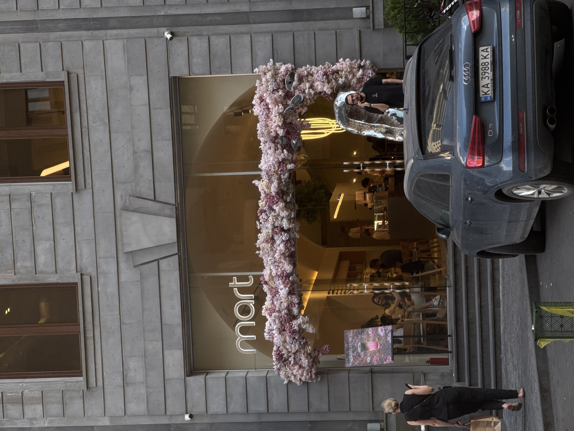 The image captures the facade of "mart" cafe in Yerevan, Armenia. The cafe's entrance is adorned with a floral arrangement. Visible inside are patrons and staff. The exterior is gray stone, with two windows on the left and a security camera mounted above the entrance.
A dark gray Audi Q3 is parked directly to the right of the entrance, partially obscuring it, with a Ukrainian license plate KA 3988 KA. A woman, dressed in black, stands on the sidewalk in front of the cafe, looking at her mobile phone while holding a shopping bag.