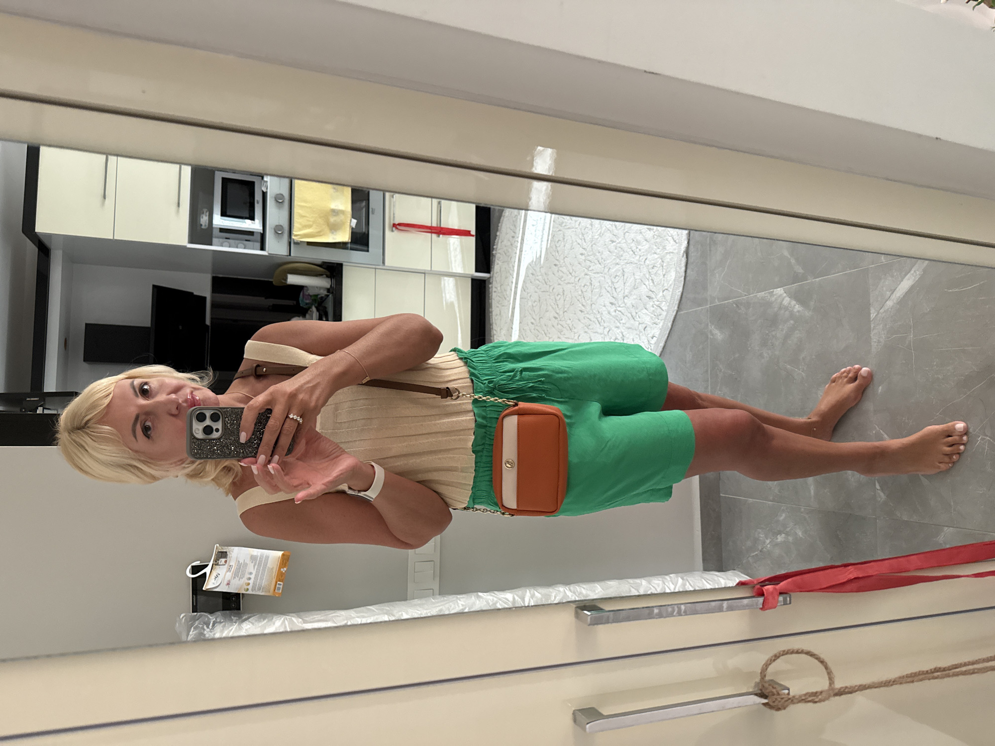The image shows a woman standing in a residential interior, likely a kitchen or living area, as indicated by the cabinetry and appliances visible in the reflection of a large mirror. The woman is taking a selfie with her phone, which is in her right hand. She has blonde hair, is wearing a light-colored ribbed tank top, and bright green shorts. A small, brown and tan crossbody bag is slung over her shoulder. Her feet are bare, and she is standing on a tiled floor. In the background, reflected in the mirror, are kitchen cabinets, a microwave, a range hood, and a stovetop. The overall environment suggests a domestic setting. There is no specific information in the image that would allow for identification of the city as Alanya, Turkey.