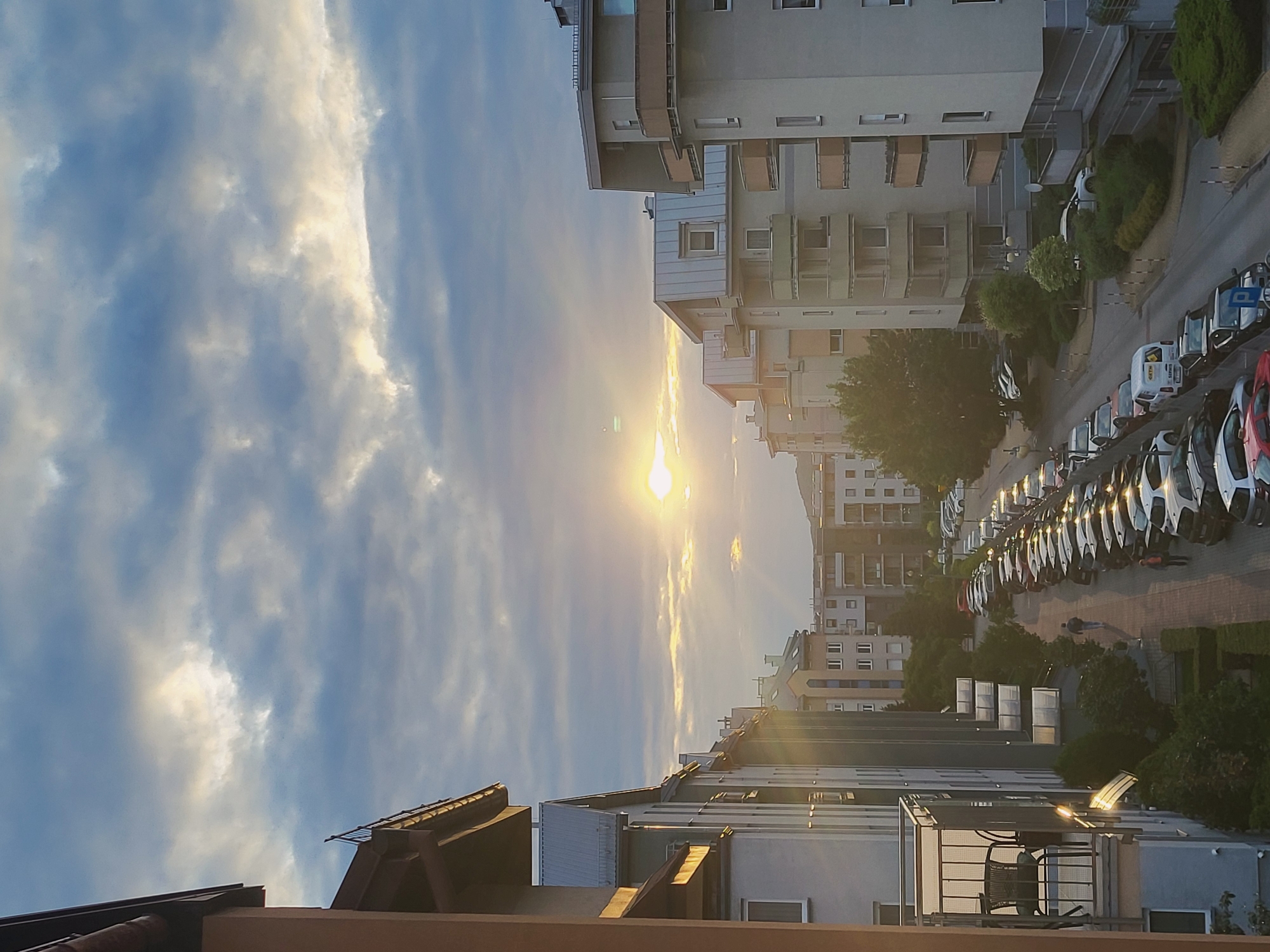 The image shows a street lined with apartment buildings, with a parked car lot running alongside the road. The sun is setting, casting a warm glow over the scene. The sky is partly cloudy, with shafts of light breaking through the clouds. A few people are walking on the sidewalk, and there are many cars parked on both sides of the street. The buildings are modern, with balconies and large windows. The street is paved, and there are trees and bushes planted along the sidewalk.