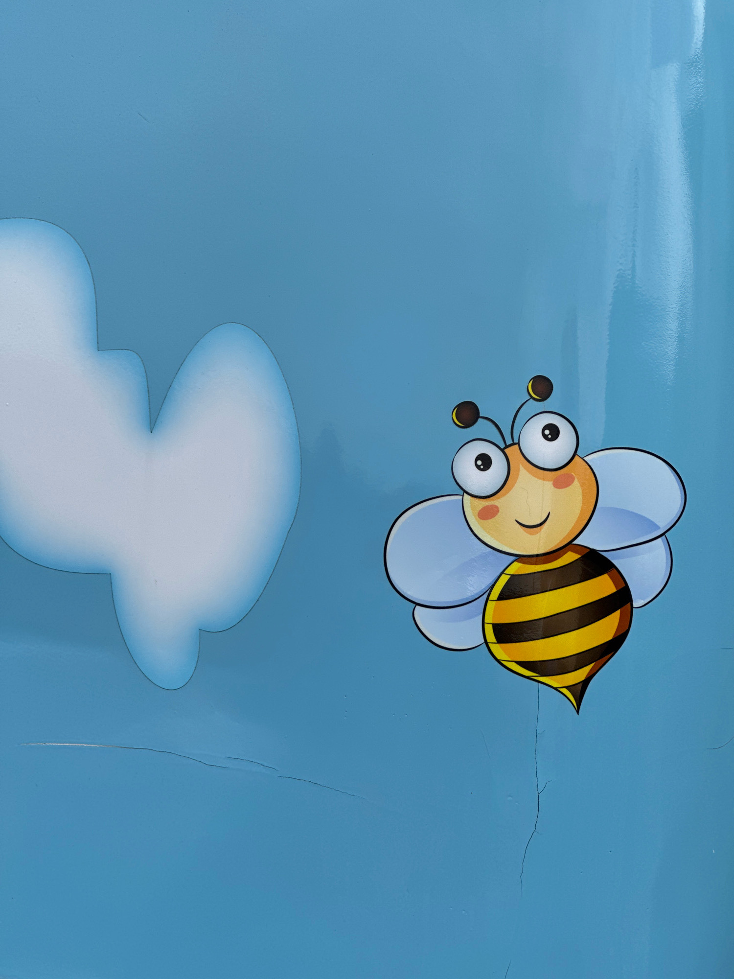 The image shows a cartoon drawing of a bee against a blue background. The bee has large, round eyes, a smiling mouth, and black and yellow stripes on its body. It has four translucent wings and two antennae with brown tips. To the left of the bee is a white, cloud-like shape with a soft, rounded outline. The blue background appears to be a wall or surface, with visible cracks and a glossy sheen in some areas. The overall impression is of a decorative element, possibly on a child's play area or a public space. The image does not contain any information that would allow for its location to be identified as Cambridge, United Kingdom.