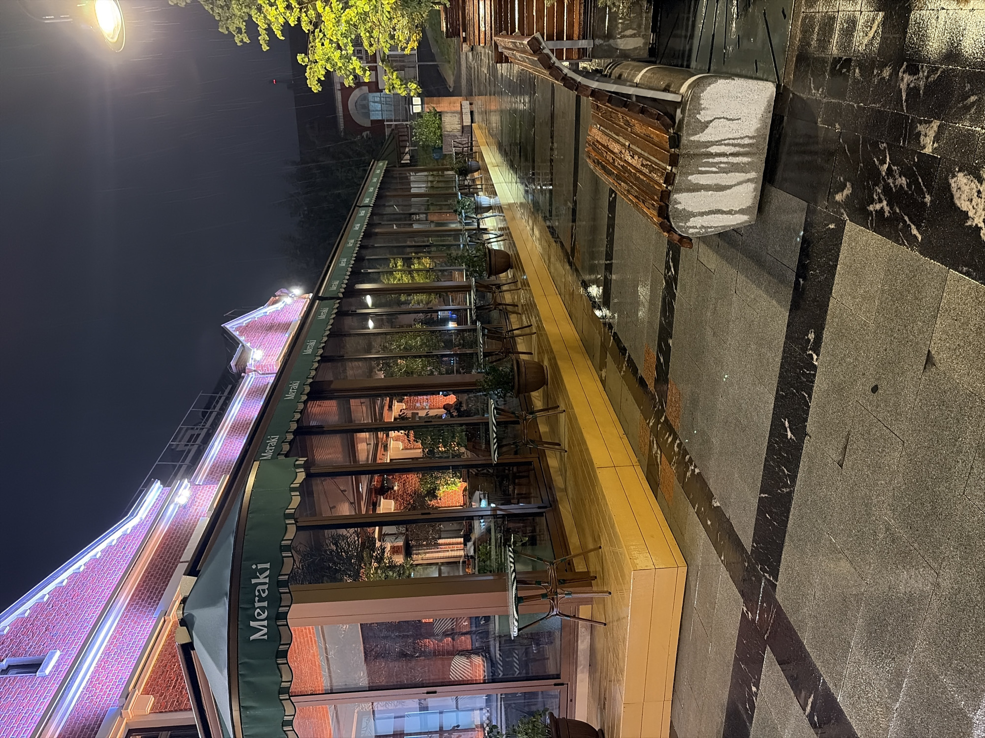 The image depicts an outdoor seating area of a restaurant at night, under heavy rain. The restaurant, identified by the "Meraki" signage on its awnings, appears to be located in Moscow, Russia, given the architectural style and the overall ambiance.
The building itself is constructed of red brick and is illuminated by soft purple and white lights along its roofline. A long, glass-enclosed dining area runs along the front of the building, with tables and chairs visible inside. Outside, there is a raised wooden platform with a few small tables and chairs, which are wet from the rain.
The ground is paved with large, dark grey and black granite tiles, interspersed with lighter grey ones. The wet surface reflects the lights, creating a glossy, almost mirror-like effect. In the foreground, a wooden bench with a stone base is positioned on the wet pavement. Water streaks are visible on the stone base of the bench, indicating the ongoing rainfall.
Further along the pavement, a series of wooden benches are arranged, also appearing wet. Trees with bright green foliage are visible to the right, their leaves illuminated by what appears to be a street lamp overhead. The sky is dark, and streaks of rain are discernible, indicating a downpour. The overall scene conveys a sense of a quiet, rainy evening at a restaurant.