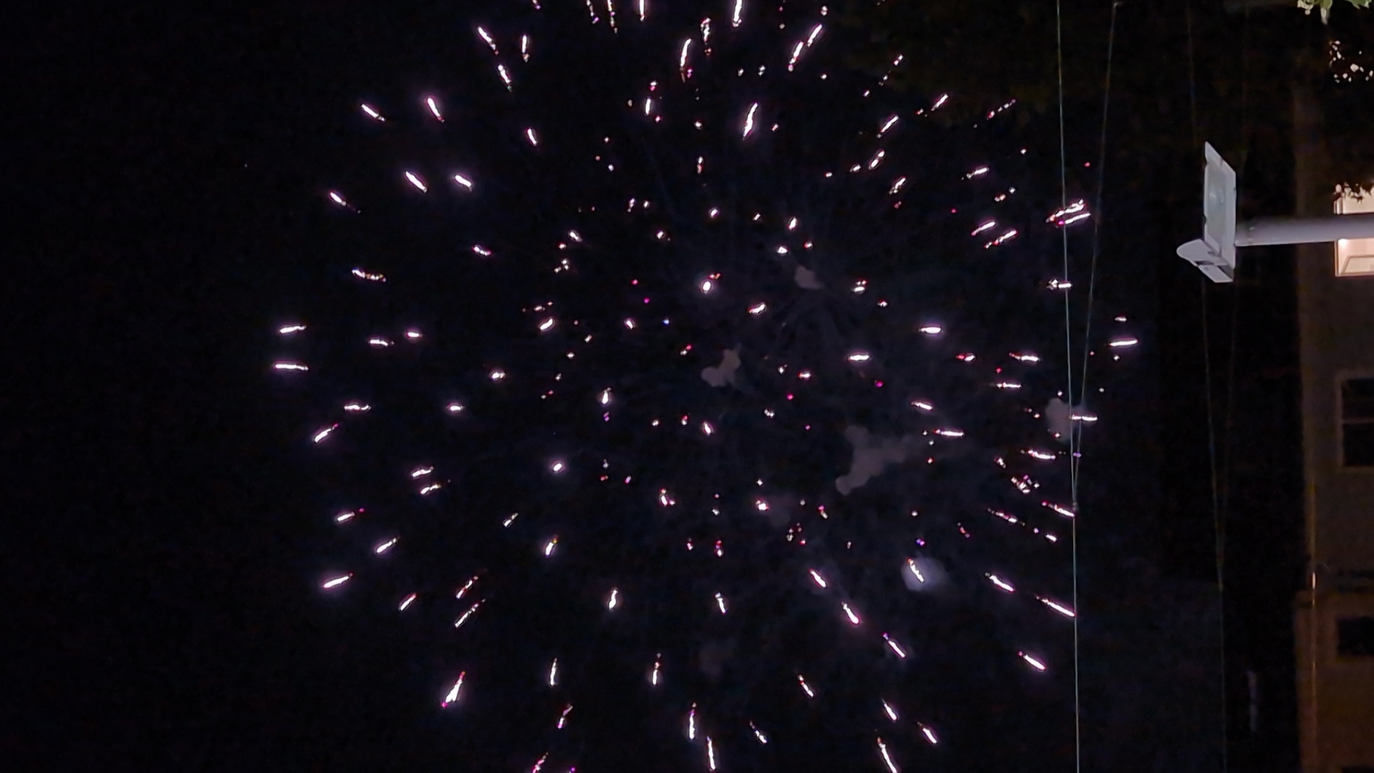 The image depicts a nighttime scene with fireworks illuminating the dark sky over the city of Bingen am Rhein, Germany. The fireworks are predominantly pink and white, appearing as streaks and bursts of light in a somewhat circular formation across the upper portion of the frame. Below the fireworks, a dark silhouette of trees and buildings is visible, with utility lines strung horizontally across the mid-ground. A white, angular structure on a pole is also present in the lower right quadrant of the image. The scene suggests a celebratory event occurring at night in an urban or semi-urban setting.