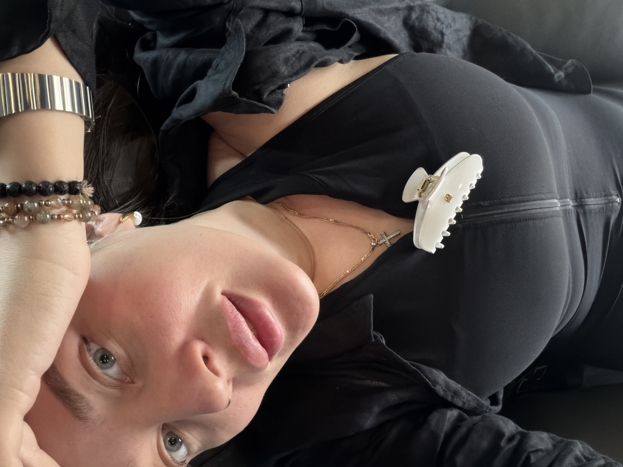 A woman lies on her back, head slightly tilted, with her left arm raised above her head and her right arm positioned to her right side. She has fair skin, light blue eyes, and pink lips. She wears multiple bracelets on her left wrist, including a metal watch, a bracelet with black beads, and another with translucent and gold beads. A small silver hoop earring is visible in her left ear. A gold necklace with a cross pendant rests on her chest. She is dressed in black clothing, including what appears to be a fitted top and a dark, possibly linen, jacket or shirt. A white hair clip is attached to the fabric near her right shoulder. The background is dark and indistinct, possibly a couch or another dark surface. The image does not contain any information indicating it was taken in Moscow, Russia.