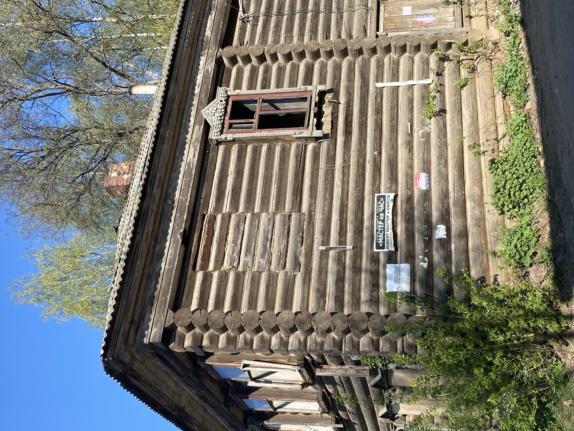 The image shows the exterior of a rustic, wooden building in Votkinsk, Russia. The building is constructed of stacked logs with decorative carvings around the window frames. One window is open, while others are boarded up. There are several advertisements and notices posted on the wooden exterior, along with some plants growing alongside the building. A section of blue sky and treetops is visible in the background.