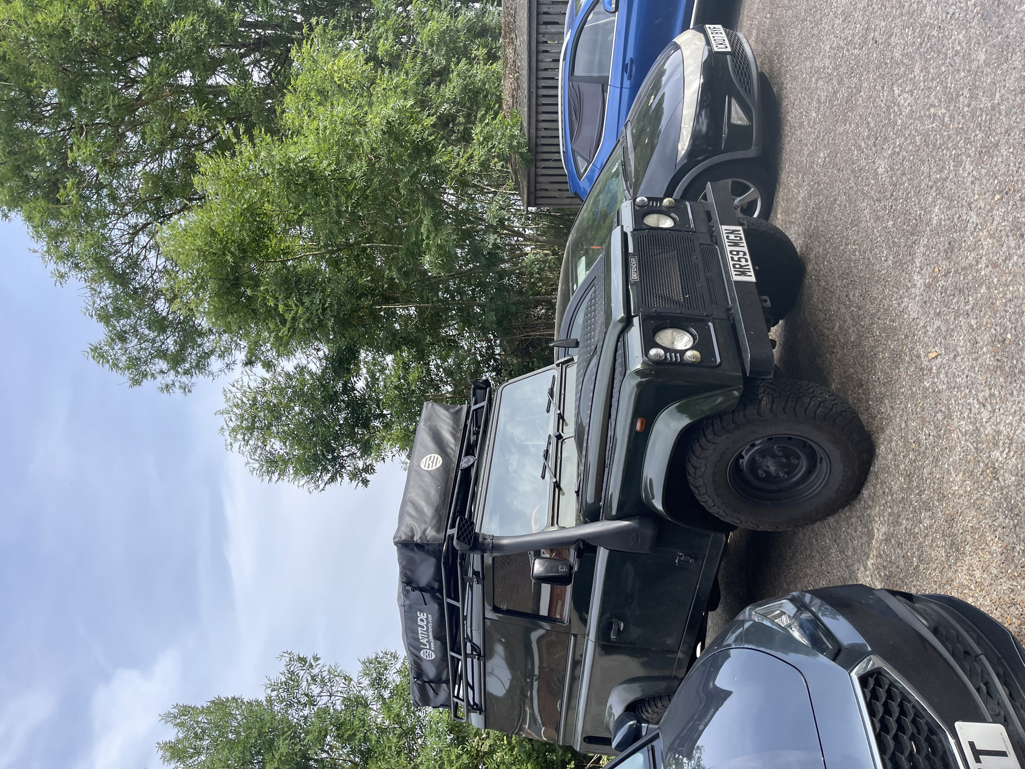 The image shows a dark green Land Rover Defender parked on an asphalt driveway in what appears to be a residential area. The vehicle has a roof rack with a tent mounted on top. The license plate visible on the front reads "MR59 MGN." Two other cars are partially visible alongside the Land Rover, one blue and the other grey. Trees and foliage are present in the background. The location is Maidstone, United Kingdom.