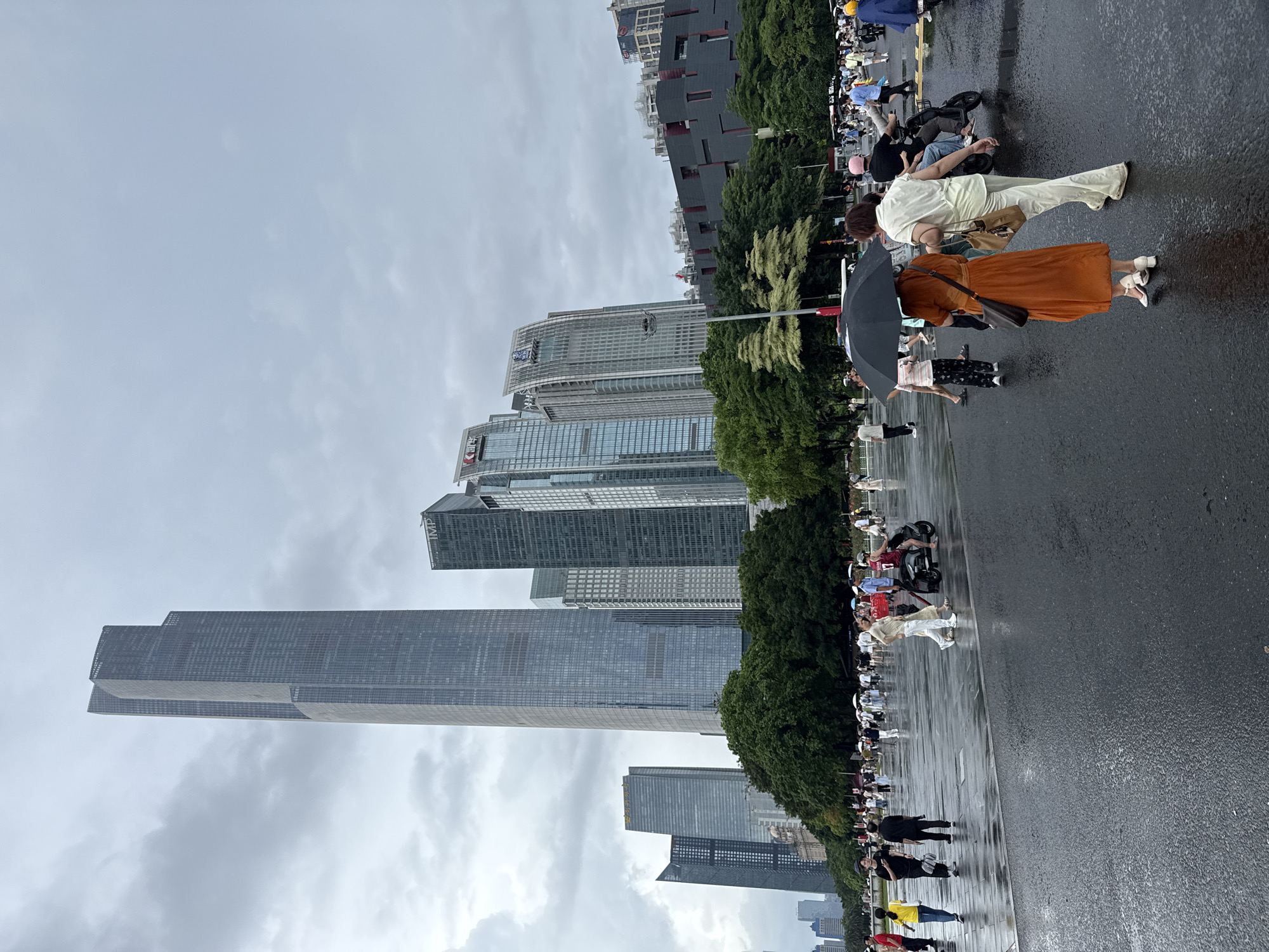 This image captures a scene in the Tianhe District of Guangzhou, China, characterized by modern skyscrapers and a public plaza. The sky is overcast, with heavy clouds suggesting potential rain. The foreground features a wet, reflective asphalt surface, indicating recent precipitation. A diverse group of people are present, some walking, others on motorcycles.
Prominent in the background are several tall, glass-facaded skyscrapers, indicative of a bustling urban center. One exceptionally tall skyscraper stands out, its sheer height dominating the skyline. Green trees form a verdant buffer between the plaza and the buildings.
In the midground and foreground, individuals are engaged in various activities. Two women are walking together, one holding a black umbrella, suggesting the possibility of rain. Another woman is riding on the back of a motorcycle. Other pedestrians are scattered across the plaza, some carrying umbrellas. The presence of multiple motorcycles and a general sense of pedestrian movement indicates a public gathering or a busy thoroughfare. The overall atmosphere is urban and contemporary, with a notable presence of architectural modernity.