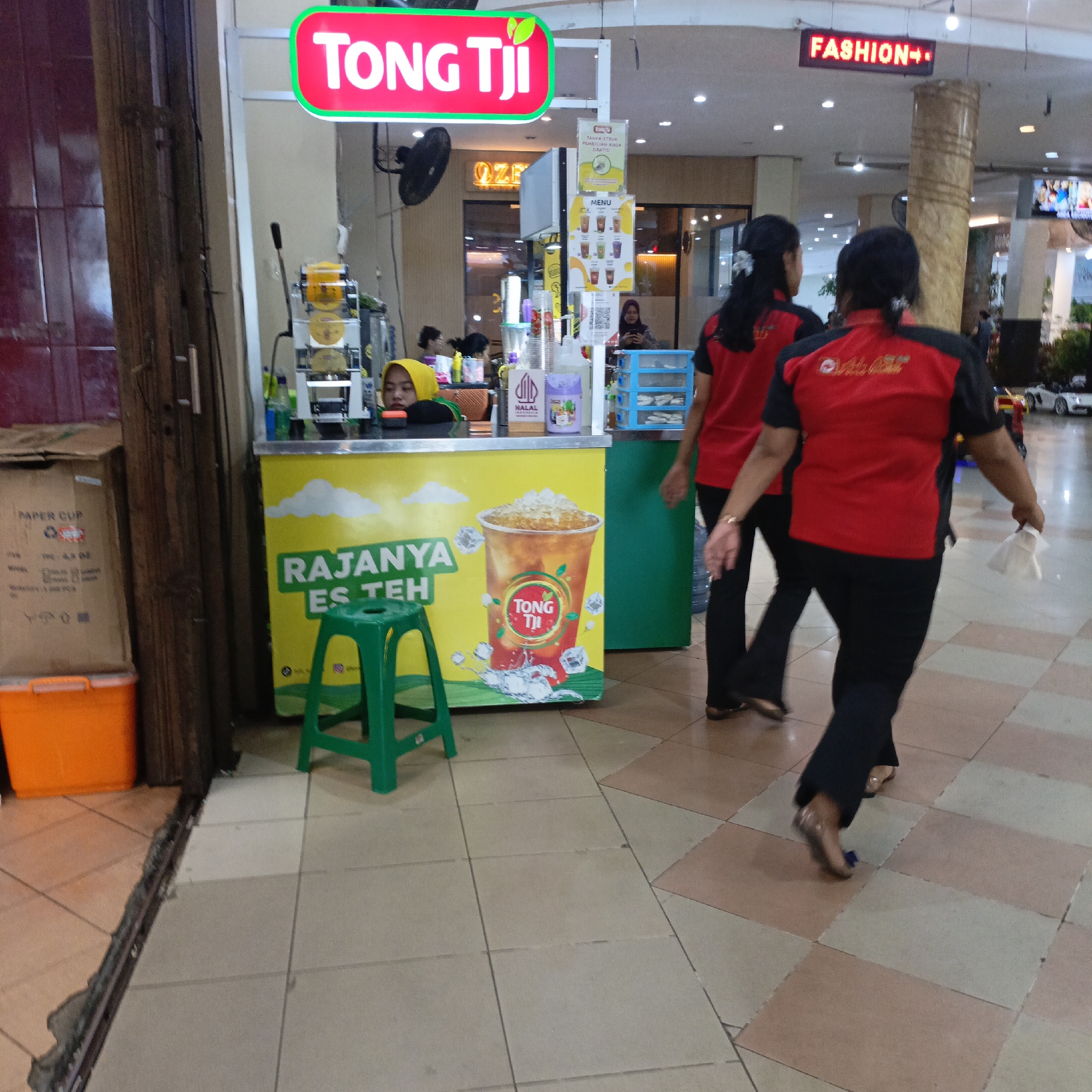 This image depicts a street-level view of a Tong Tji tea stall within a commercial area in Salatiga, Indonesia. The stall is positioned under a bright, pink "Tong Tji" sign with a green border, indicating a franchise establishment. The stall itself is predominantly yellow with white cloud graphics and a large image of an iced tea drink. Text on the stall reads "RAJANYA ES TEH" (King of Iced Tea). A young woman wearing a yellow hijab is visible behind the counter, appearing to be an employee. Adjacent to the stall, a green plastic stool is placed on the tiled floor. To the left of the stall, a cardboard box labeled "PAPER CUP" and an orange plastic bin are stacked. In the background, other shop fronts and shoppers are partially visible. A "FASHION" sign is illuminated above a pillar, suggesting the presence of a clothing store. Two women in matching red and black uniforms, likely employees of another establishment, are walking away from the camera. The floor is tiled in a checkered pattern of beige and pale pink squares. The overall environment appears to be an indoor market or a shopping center arcade.
