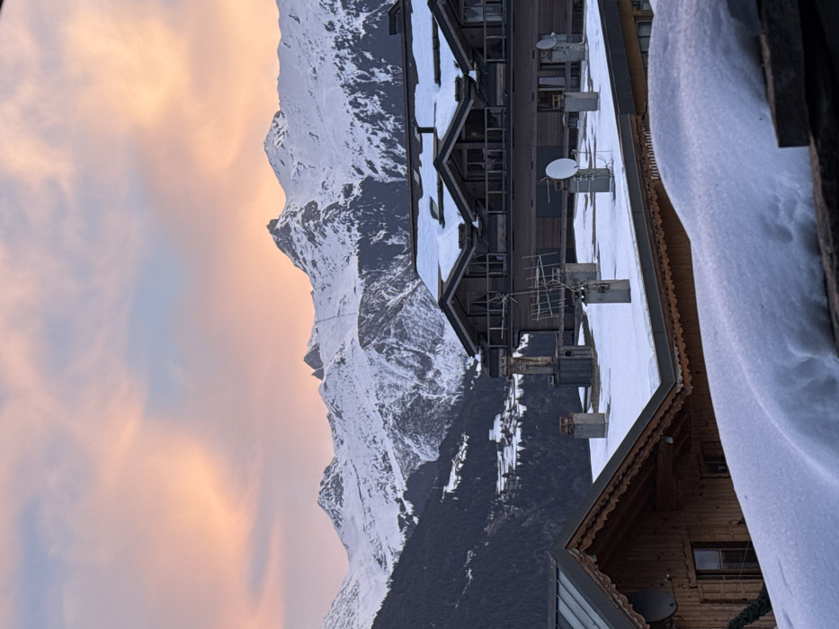 A picturesque scene showcases snow-covered mountains against a sunset-tinged sky. To the right, a building with multiple satellite dishes and antennas is partially covered in snow. The architecture exhibits a unique angled roof. The scene is likely captured from inside another building, evidenced by the wood detail and snow-covered edge on the right. The overall ambiance is serene and cold.