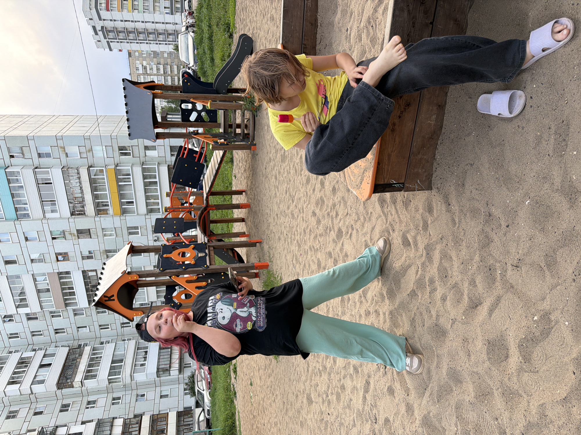 Two children are at a playground next to apartment buildings. One child is wearing a yellow t-shirt and sitting on a wooden bench, holding a popsicle. The other child is wearing a black t-shirt and light green pants, standing in the sand. The playground is located in the city of Chaikovsky, Russia.