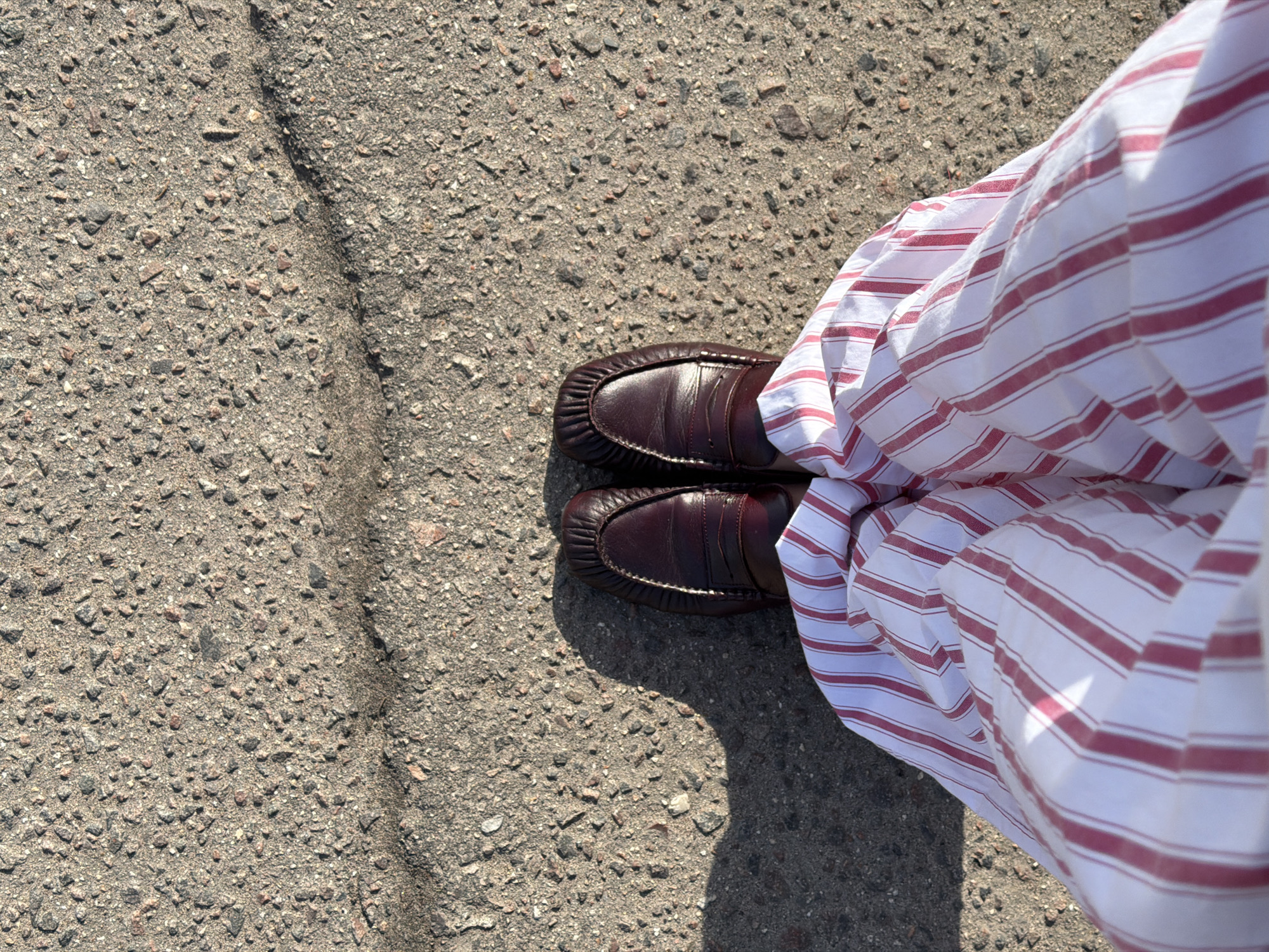 The image shows a pair of brown leather loafers on a cracked asphalt surface. A white and red striped fabric, possibly a skirt or pants, is draped over the right side of the image, partially obscuring the shoes. The asphalt surface is predominantly gray with small pebbles and a visible crack running across the upper left quadrant of the image. The overall lighting suggests a sunny day. There is no information available about the location being Mishutkino, Russia.