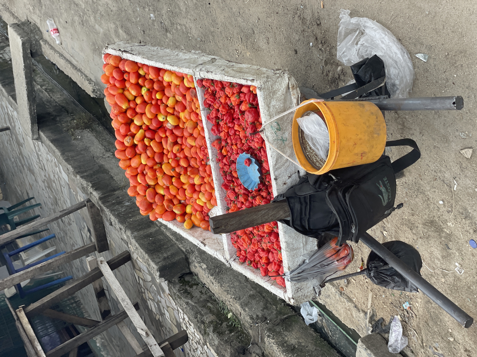 Scene: A street-side vendor in Ado, Nigeria displays tomatoes and scotch bonnet peppers in a white, divided container.
Subjects: Tomatoes (mostly ripe, orange-red), scotch bonnet peppers (red), a bucket, a black backpack with a Lacoste logo, a metal stand, trash bags, a plastic bag containing metal parts, an umbrella, and a blue plastic measuring cup.
Environment: Concrete ground and walls, a stream or drainage ditch alongside the vendor's setup, discarded furniture (chairs, wood).
Objects: Container divided into two sections, holding tomatoes in one half and scotch bonnet peppers in the other.
Actions/Interactions: The vendor has arranged the tomatoes and peppers for sale. The bucket, backpack, stand, and other items are likely part of the vendor's setup for transporting and selling the produce.
People: No people are visible in the image.