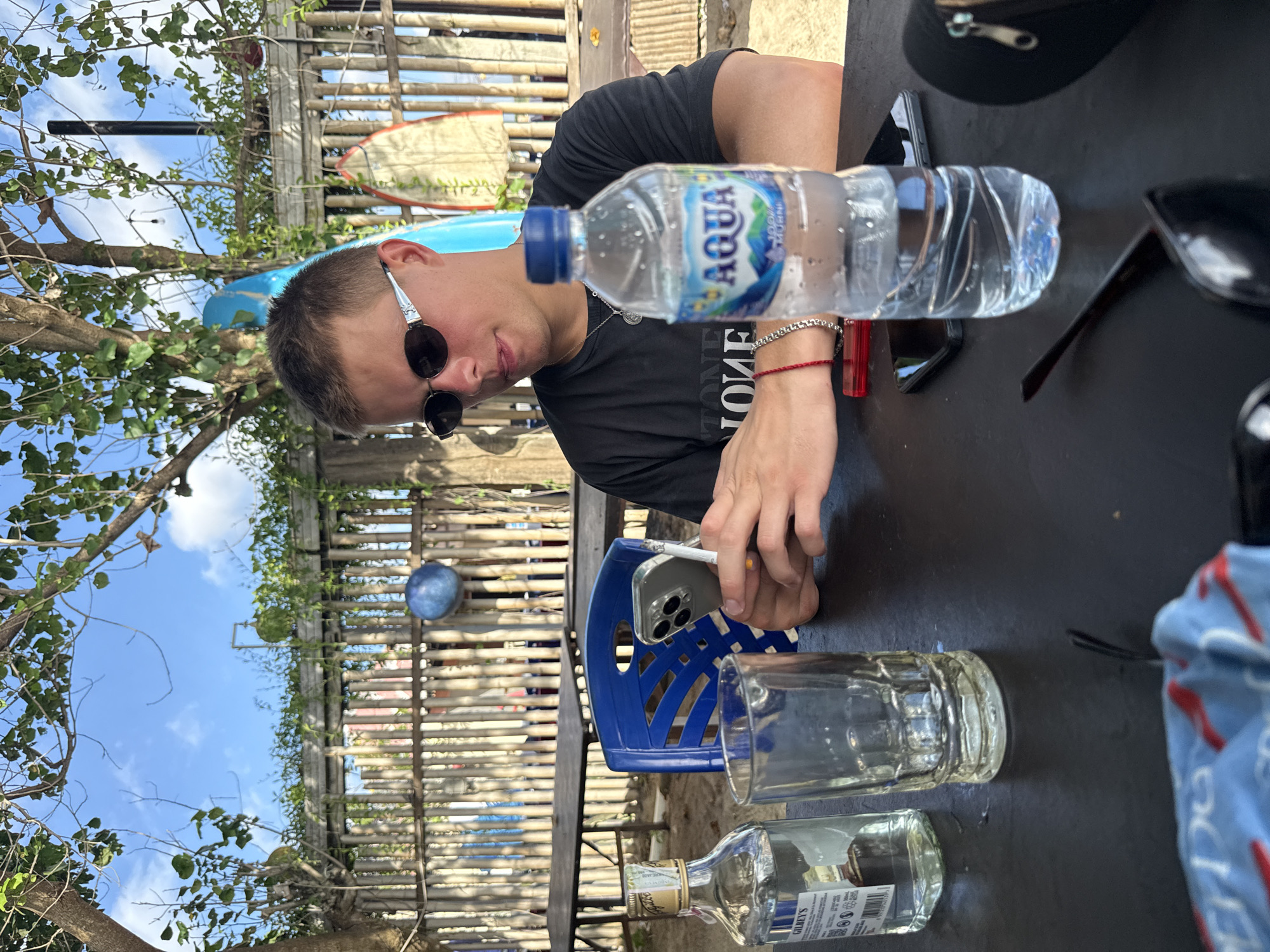 A young man sits at an outdoor table in Kuta, Indonesia. He wears sunglasses and a black t-shirt, and has a water bottle and smartphone on the table. A plastic chair is visible, along with empty glasses and a liquor bottle, suggesting a relaxed, casual setting. In the background, there's a bamboo fence, blue sky, and trees, adding to the outdoor ambiance.